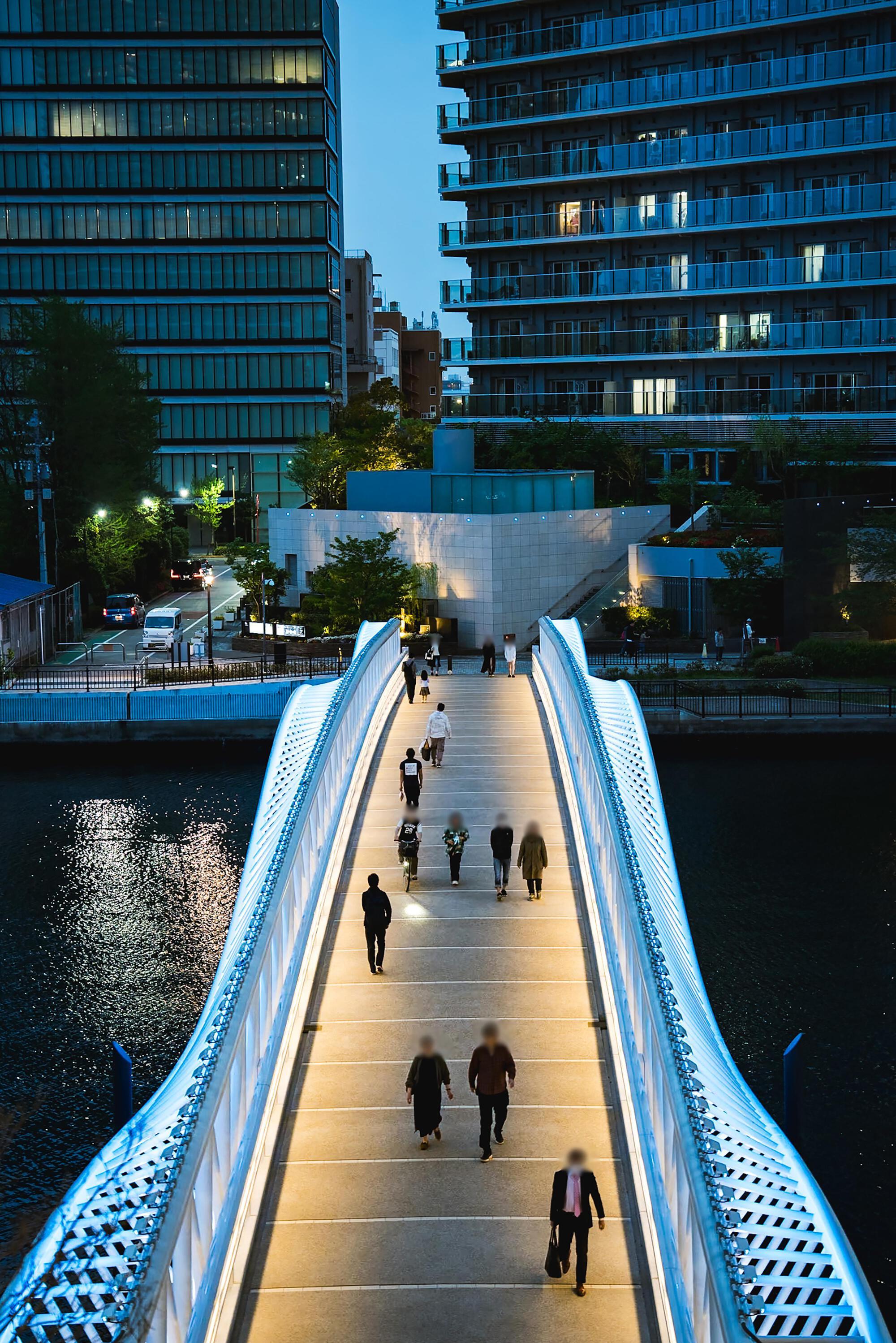 Illuminated pedestrian bridge with perforated steel railings spanning a canal at dusk with people walking