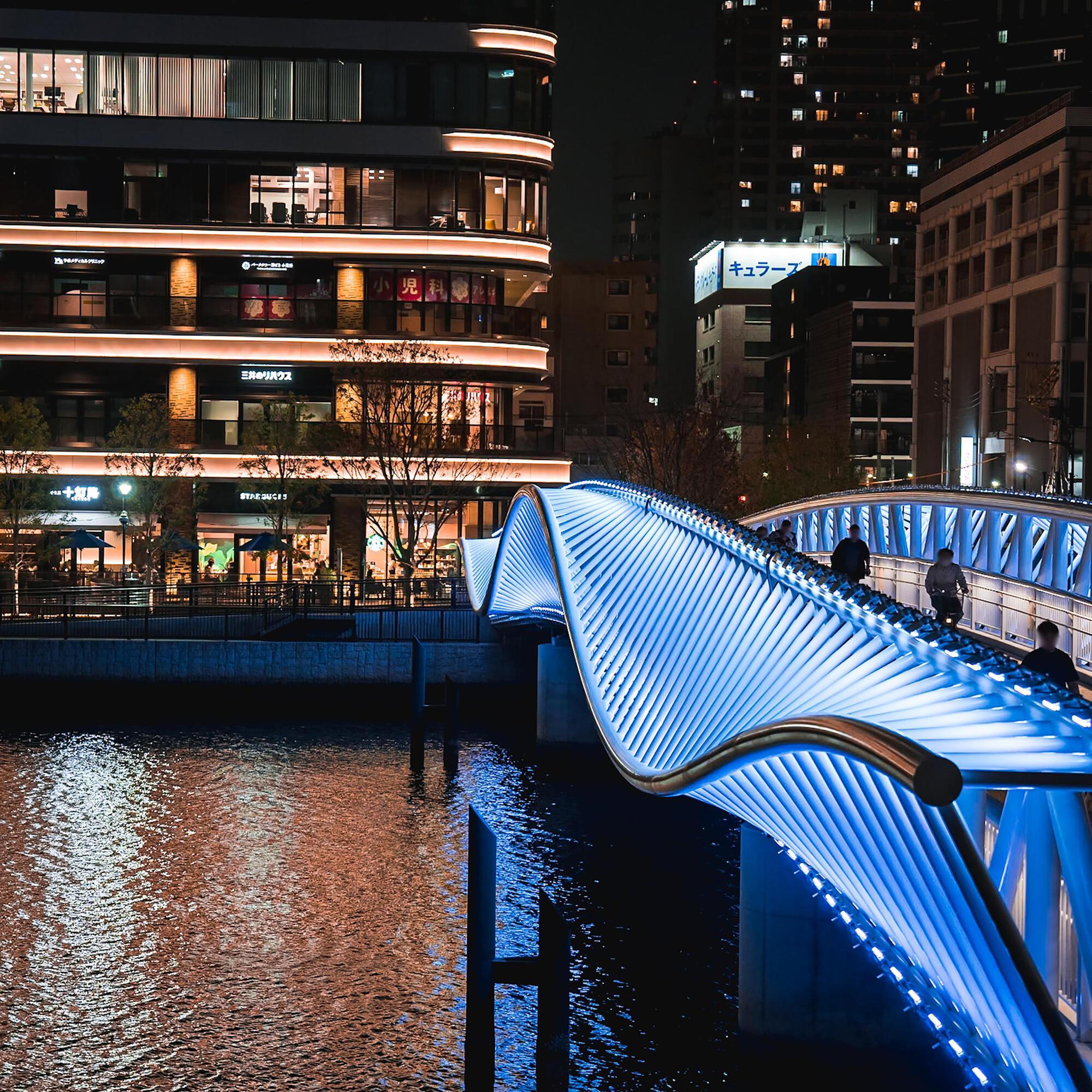 Undulating footbridge with LED-lit ribbed structure crossing dark water beside a commercial building at night