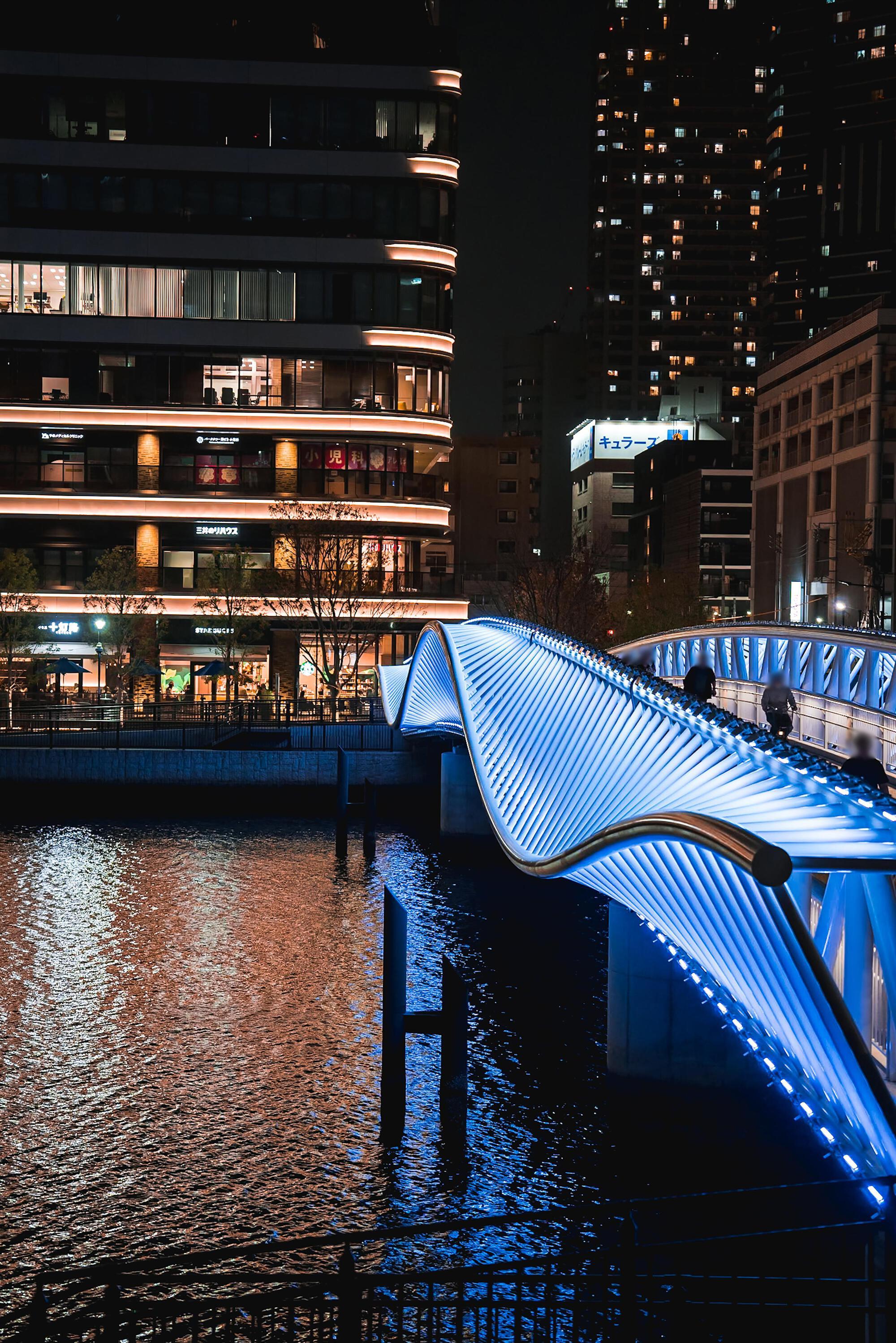 Curved pedestrian bridge with blue illuminated pleated metal canopy reflected in the canal below