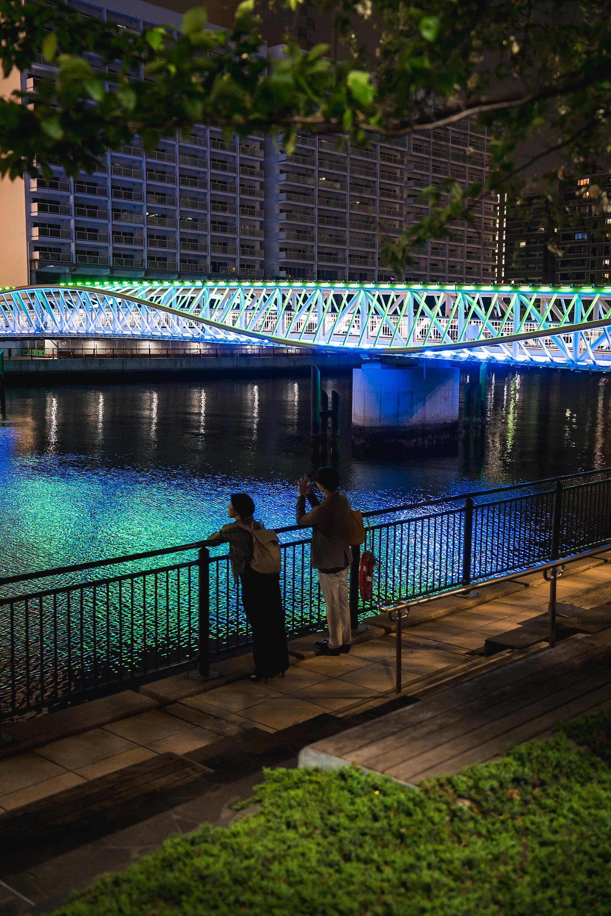 Two figures photographing the lit truss bridge from a waterfront promenade framed by foliage