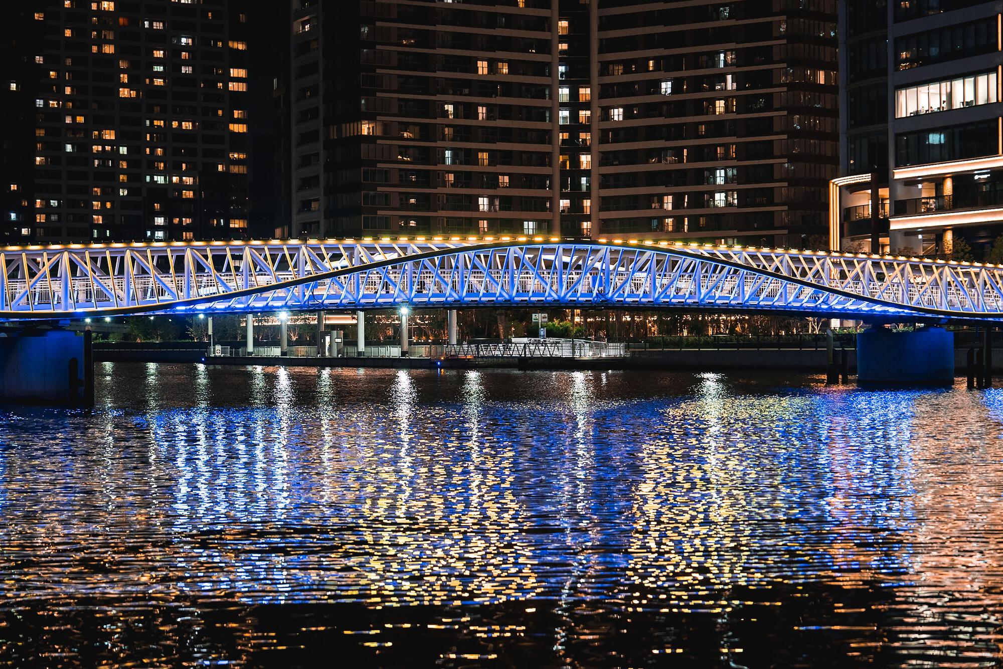 Blue-illuminated pedestrian bridge with lattice structure spanning a canal at night, reflecting in the water