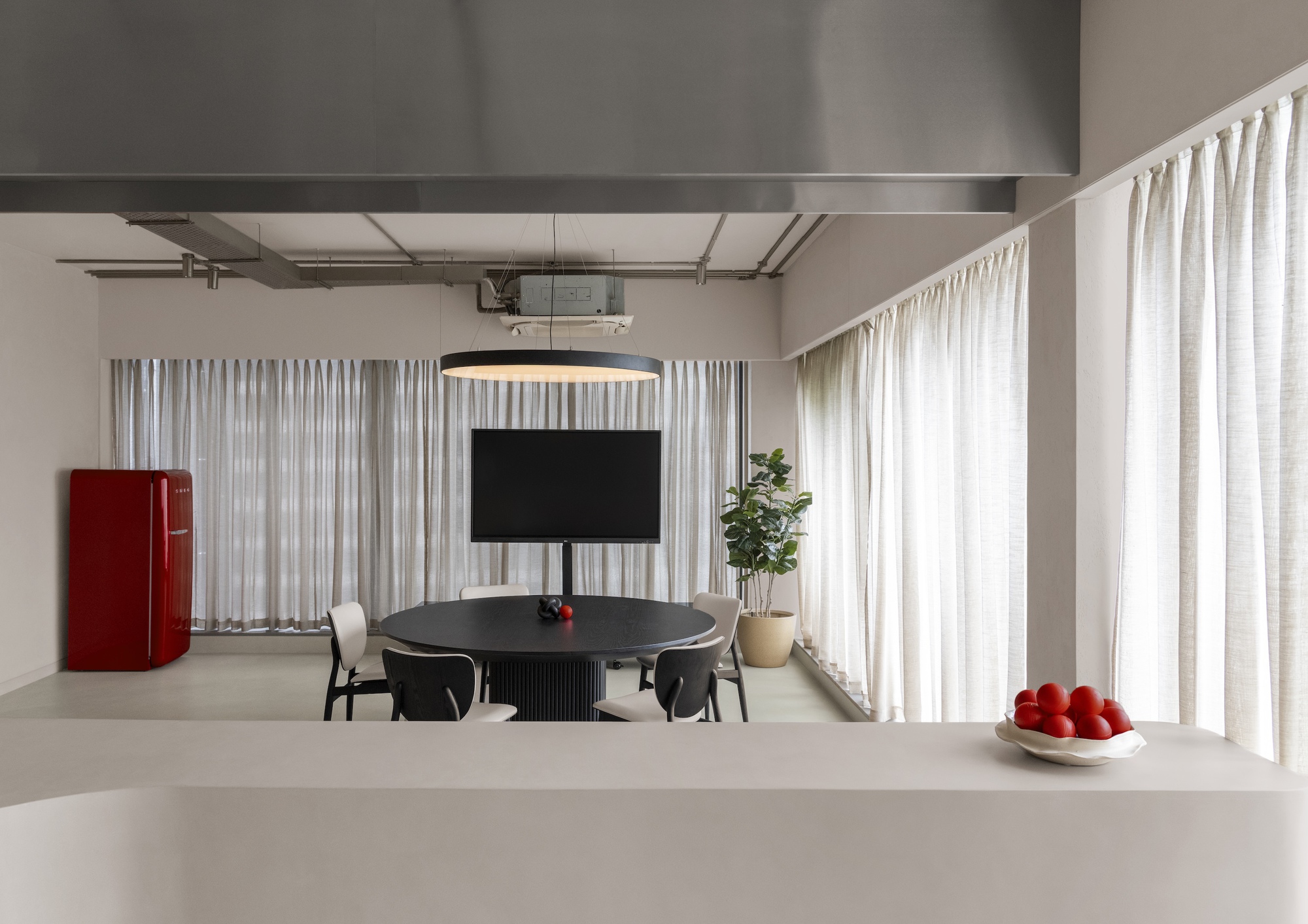 Open plan dining space with white curtains, exposed ceiling ducts, and a red refrigerator at the perimeter