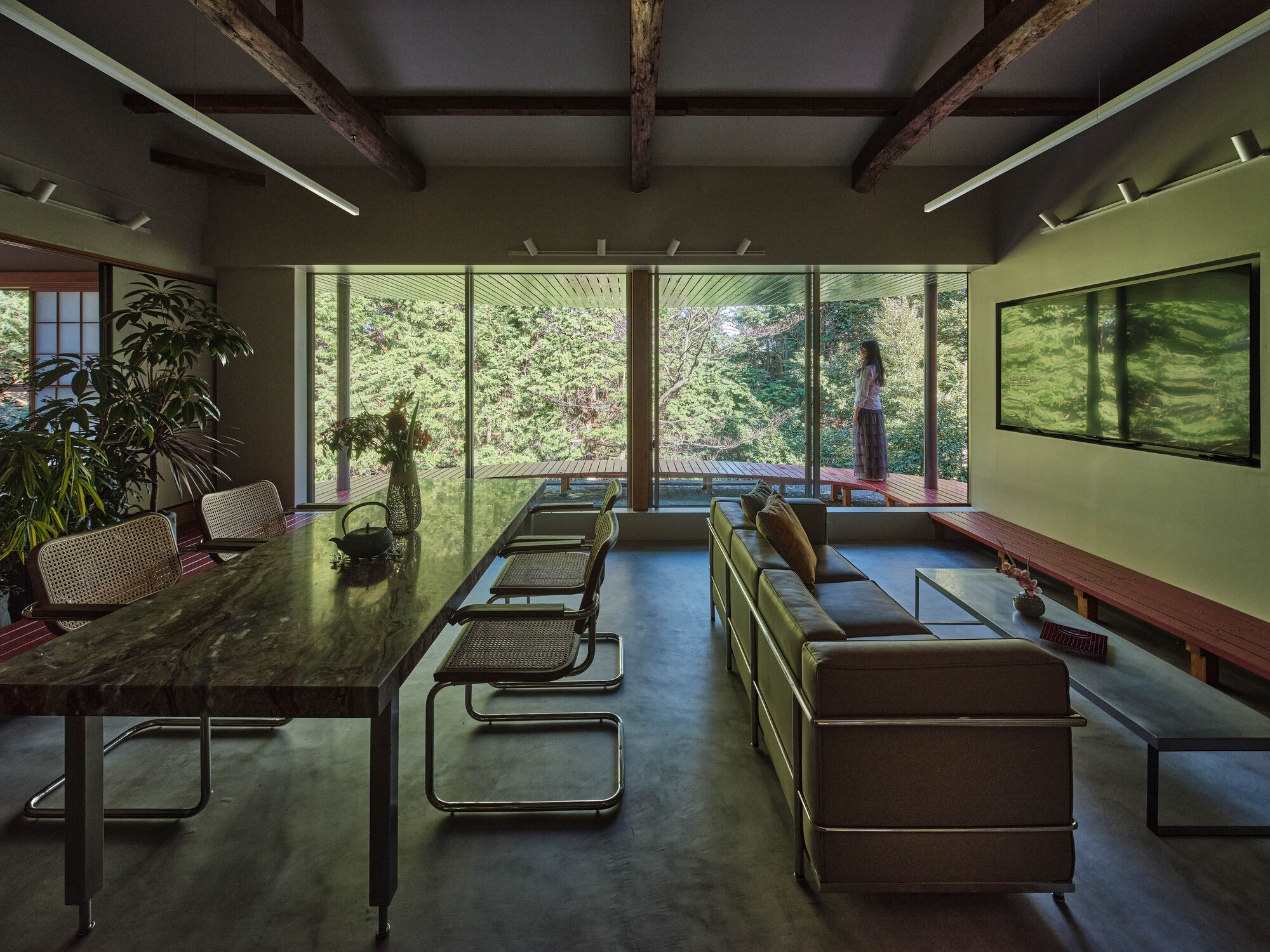 Interior living space with exposed timber beams and floor-to-ceiling glazing overlooking garden with figure standing outside