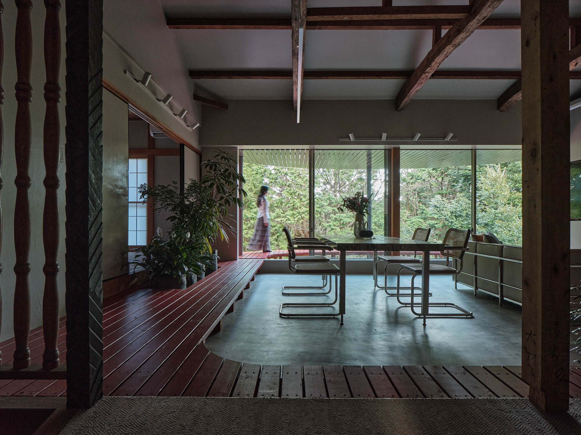 View through timber columns toward dining space with figure walking past glazed wall and garden beyond