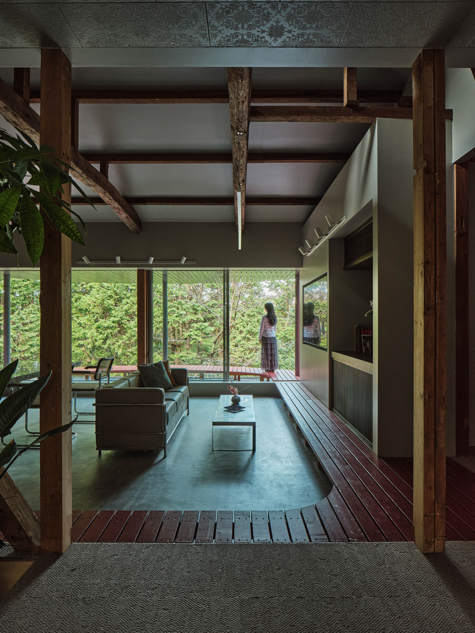 Living room with exposed timber frame and glass wall overlooking garden with figure standing outside