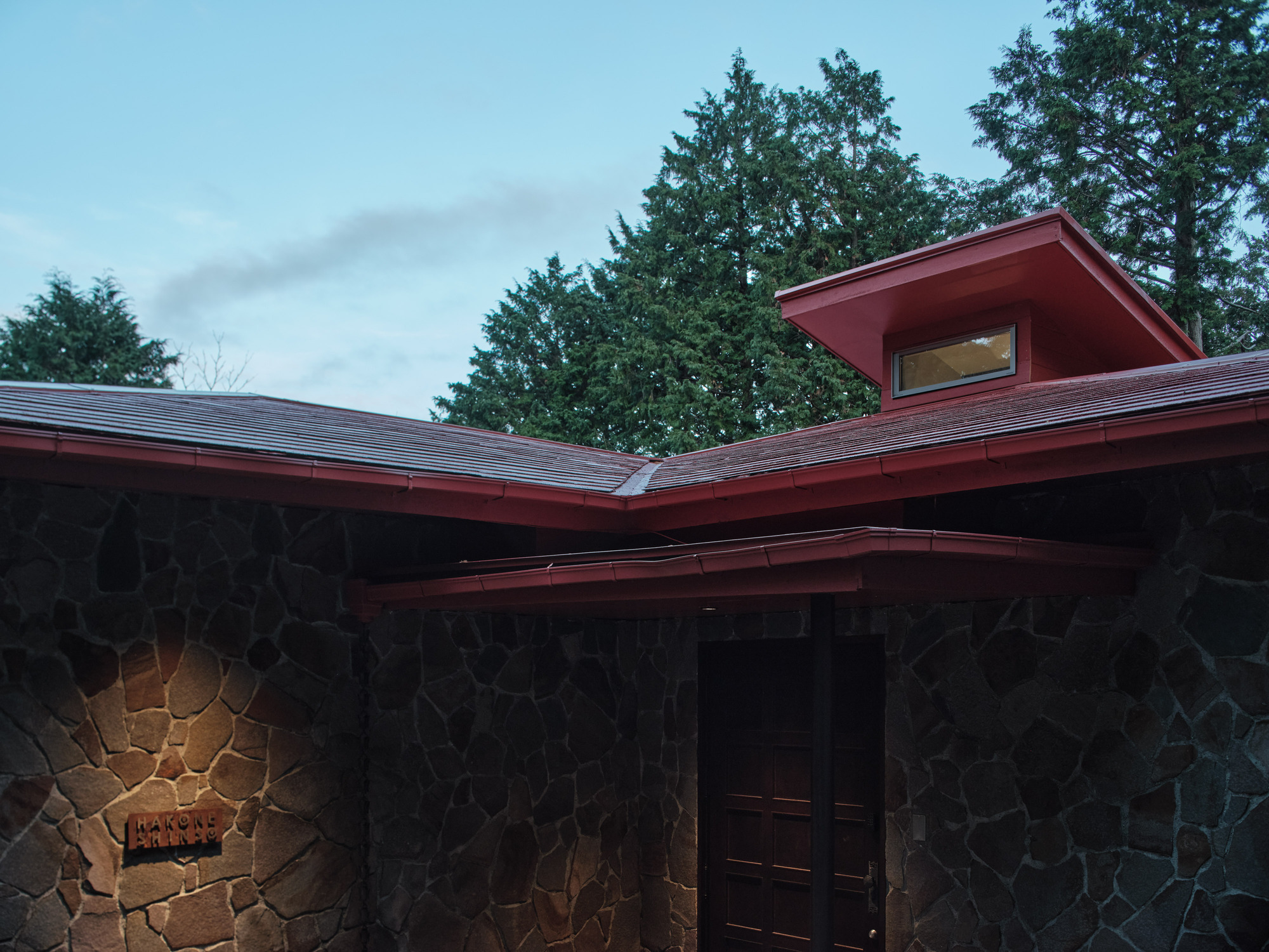 Angled roofline with red eaves and clerestory window set among tall evergreen trees at dusk