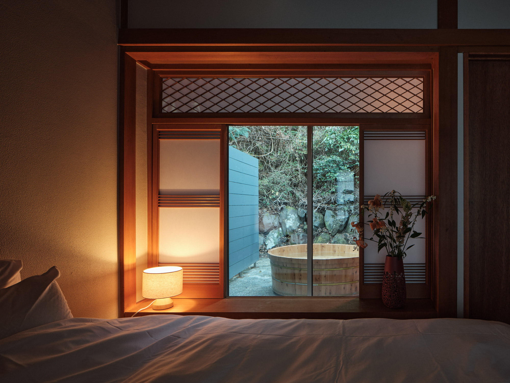 Bedroom view through sliding doors to outdoor soaking tub framed by wooden lattice and a glowing floor lamp