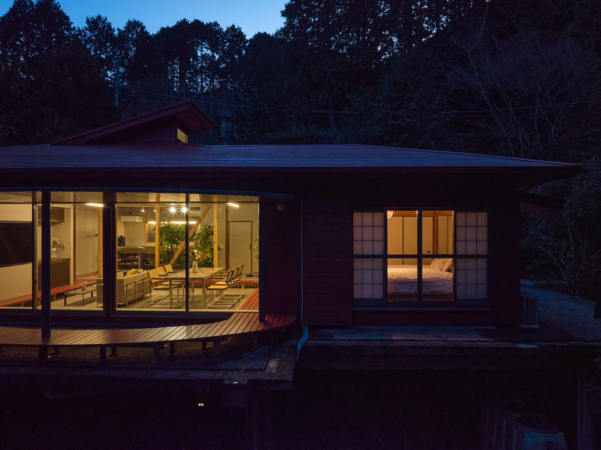 Illuminated living pavilion with glazed walls and timber deck at nightfall beside a dark reflecting pond
