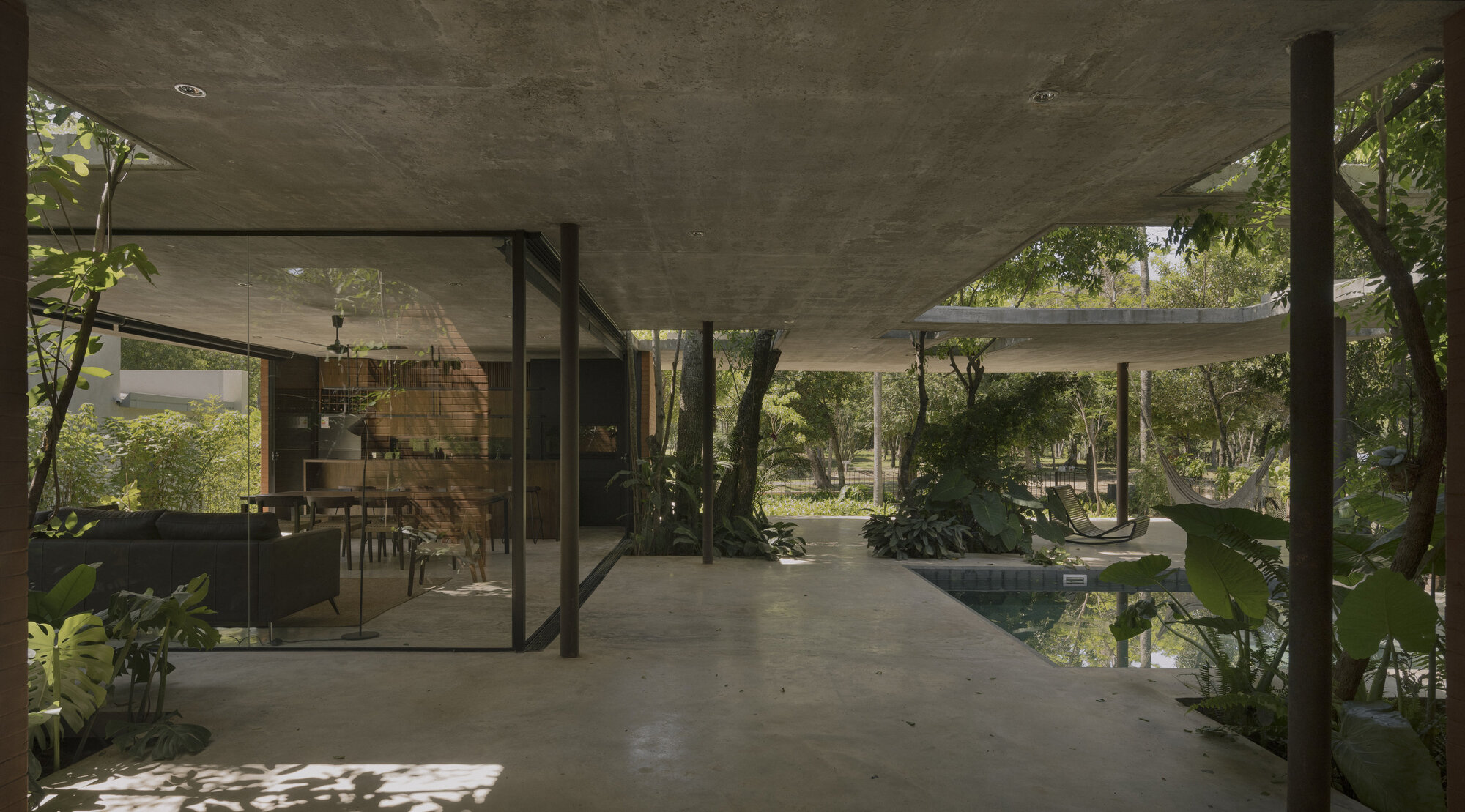 Open pavilion with concrete ceiling connecting glazed dining room to water garden and wooded landscape beyond