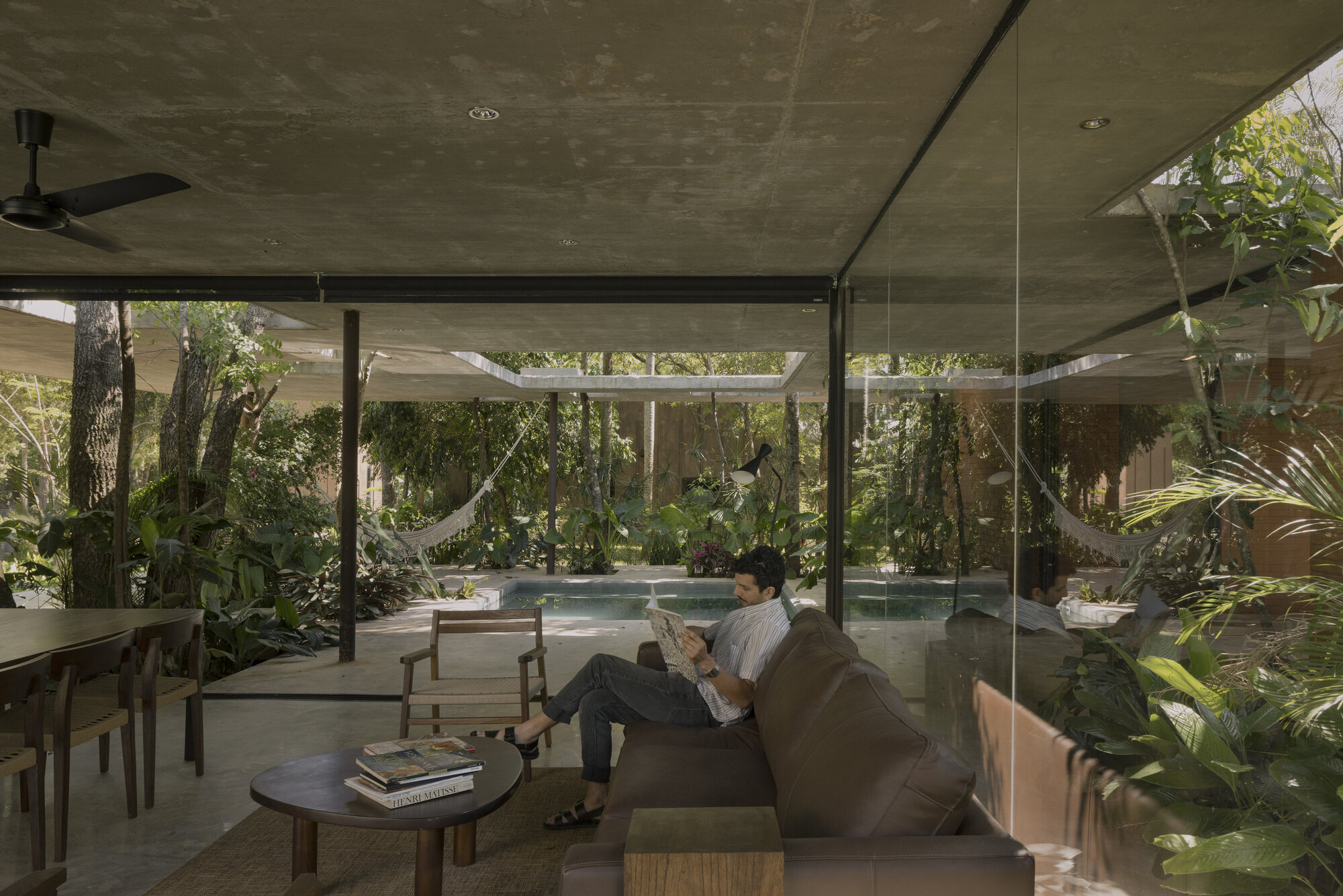 Interior living space with person reading on sofa overlooking pool and garden through floor-to-ceiling glass walls