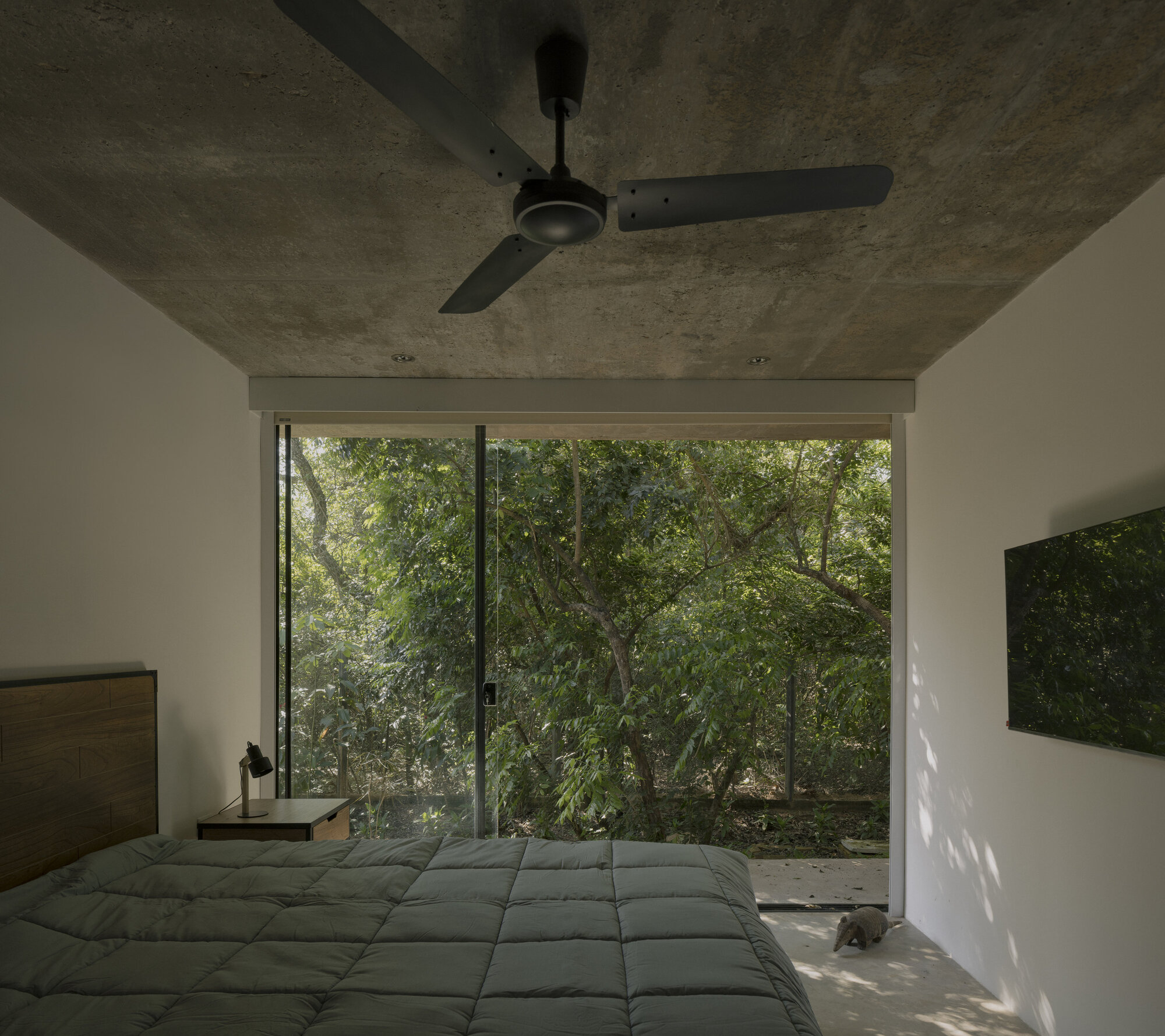 Bedroom with exposed concrete ceiling and ceiling fan facing full-height glazing opening to forested landscape