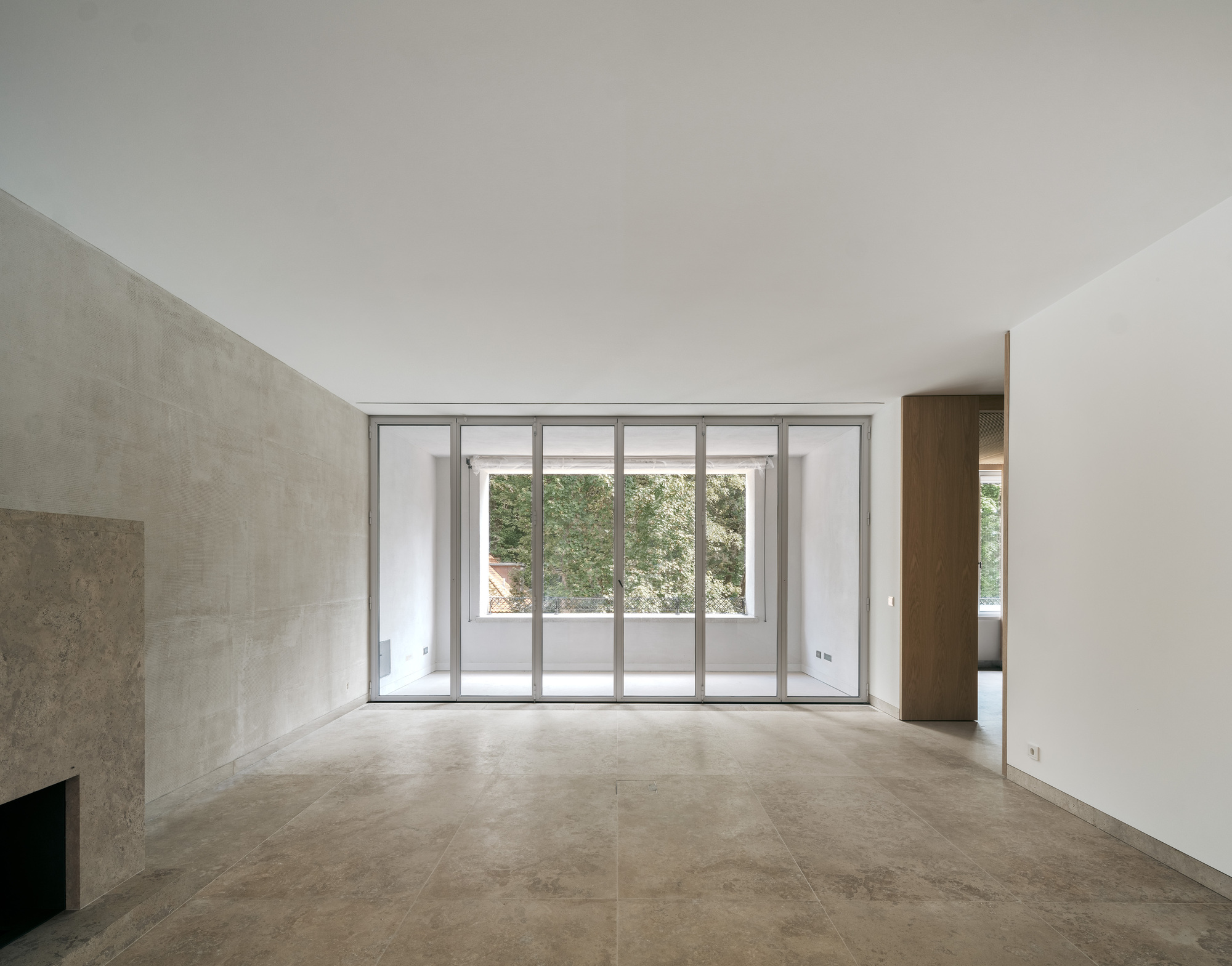 Empty room with large stone floor tiles and sliding glass doors overlooking a terrace with trees beyond