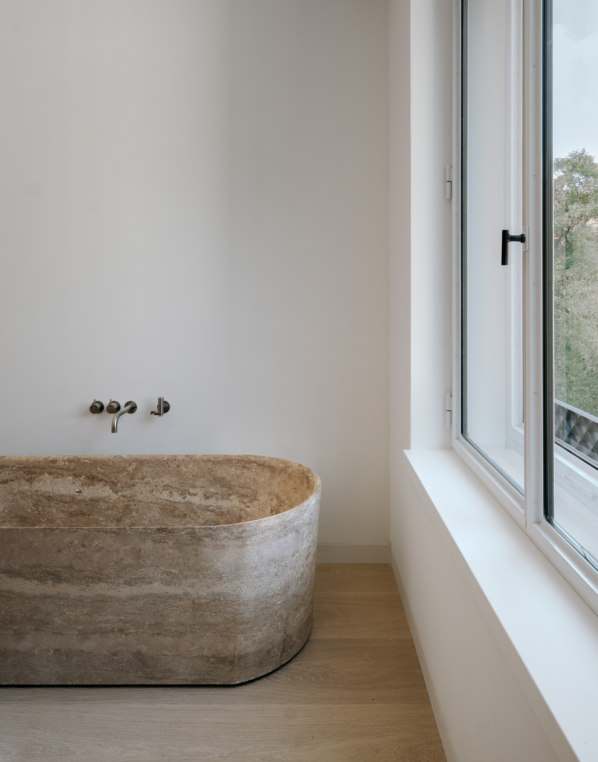 Freestanding concrete bathtub beneath wall-mounted faucets next to a narrow casement window with treetops visible outside