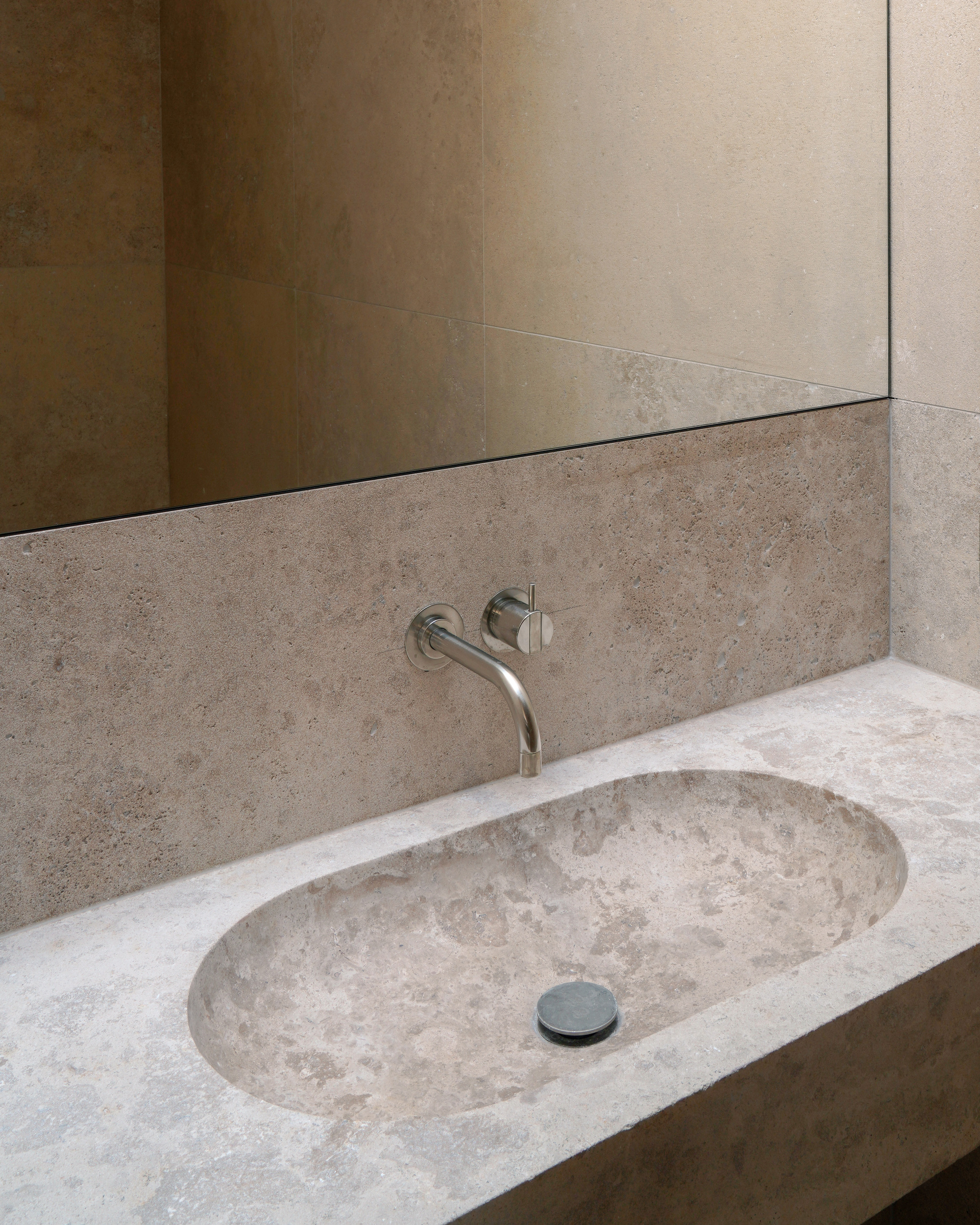 Travertine vanity with integrated oval sink and wall-mounted faucet beneath a frameless mirror