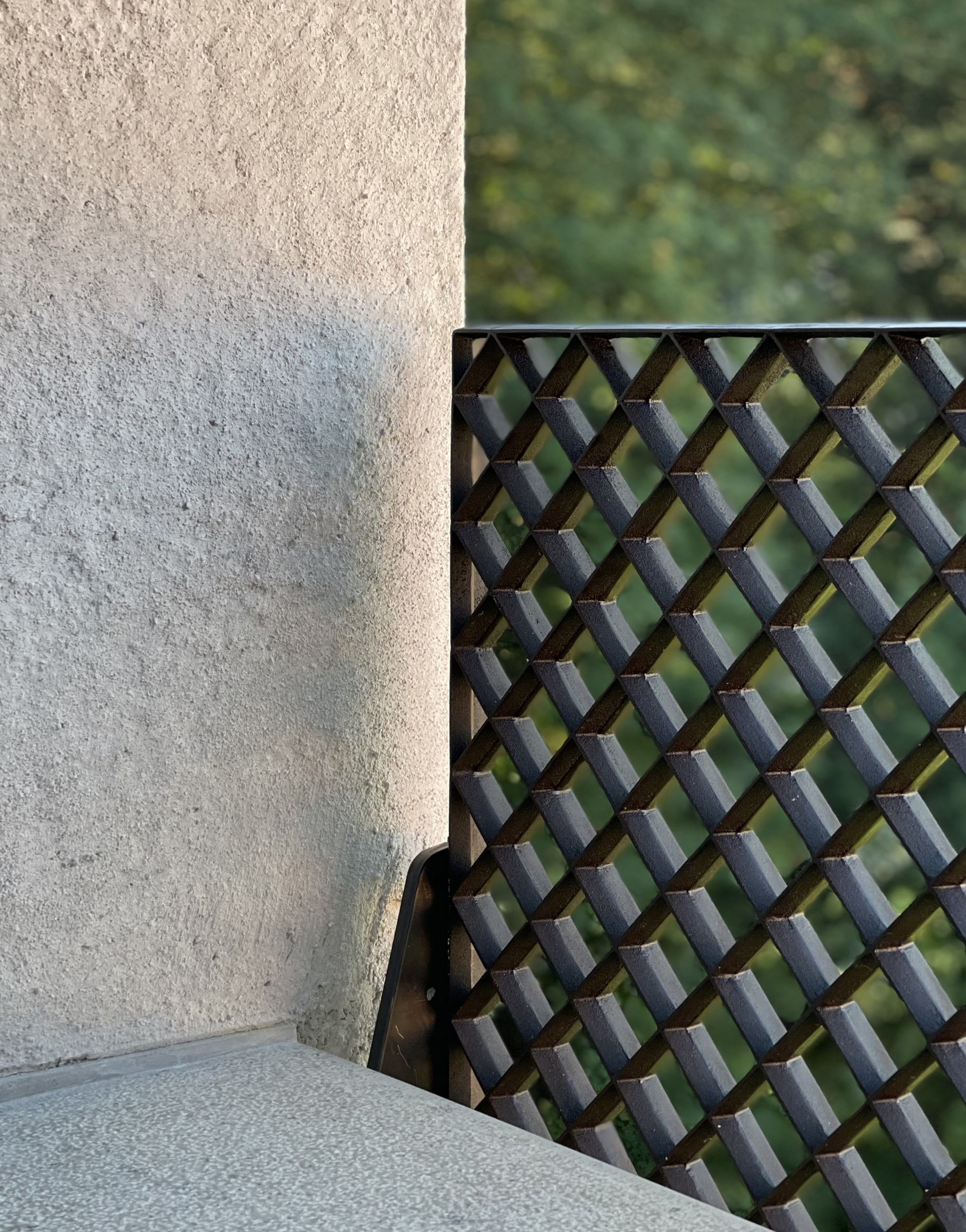 Woven metal screen beside textured plaster wall with blurred foliage beyond