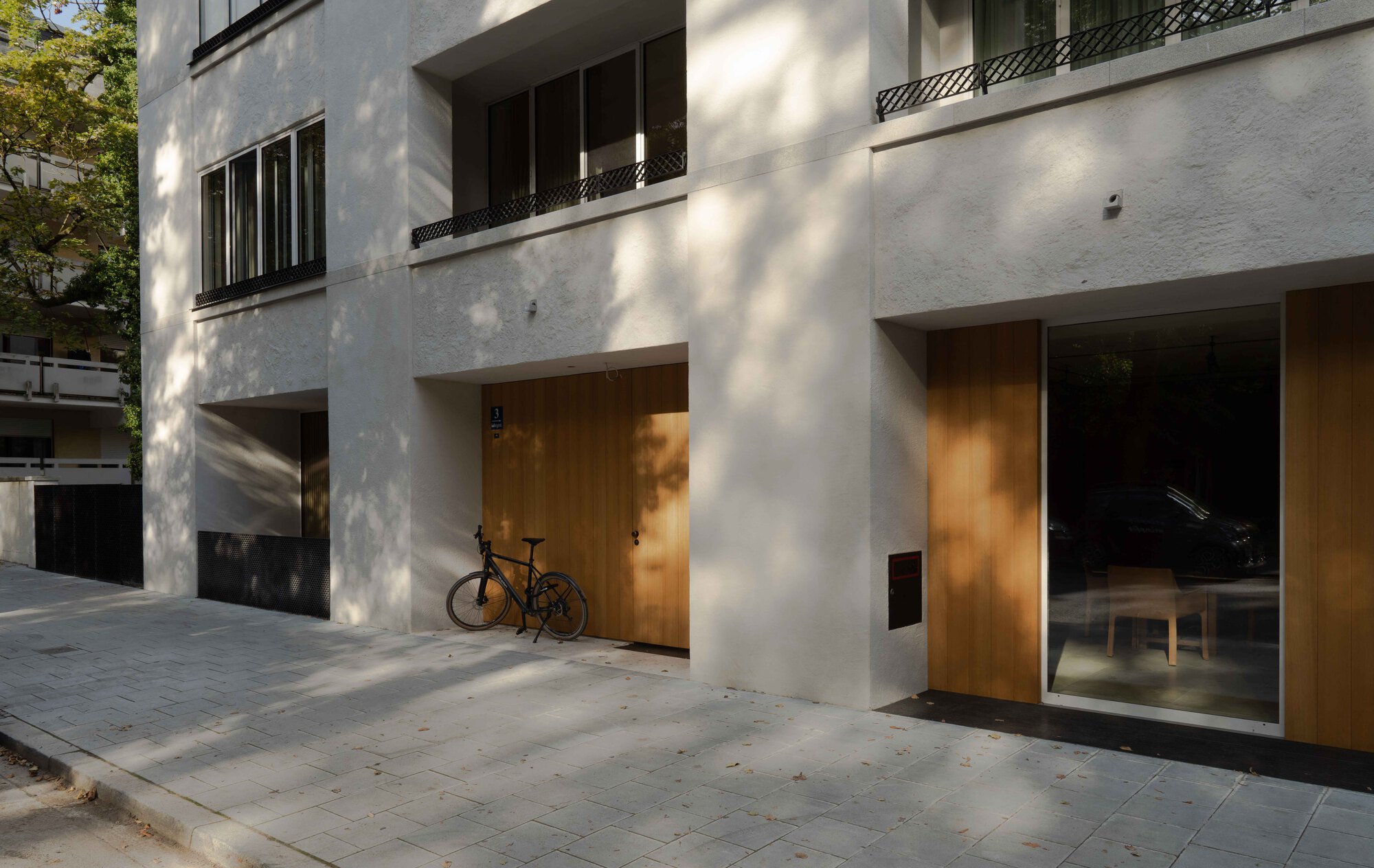 Street-level facade with timber-clad entrances and glass storefronts beneath dappled tree shade with a bicycle