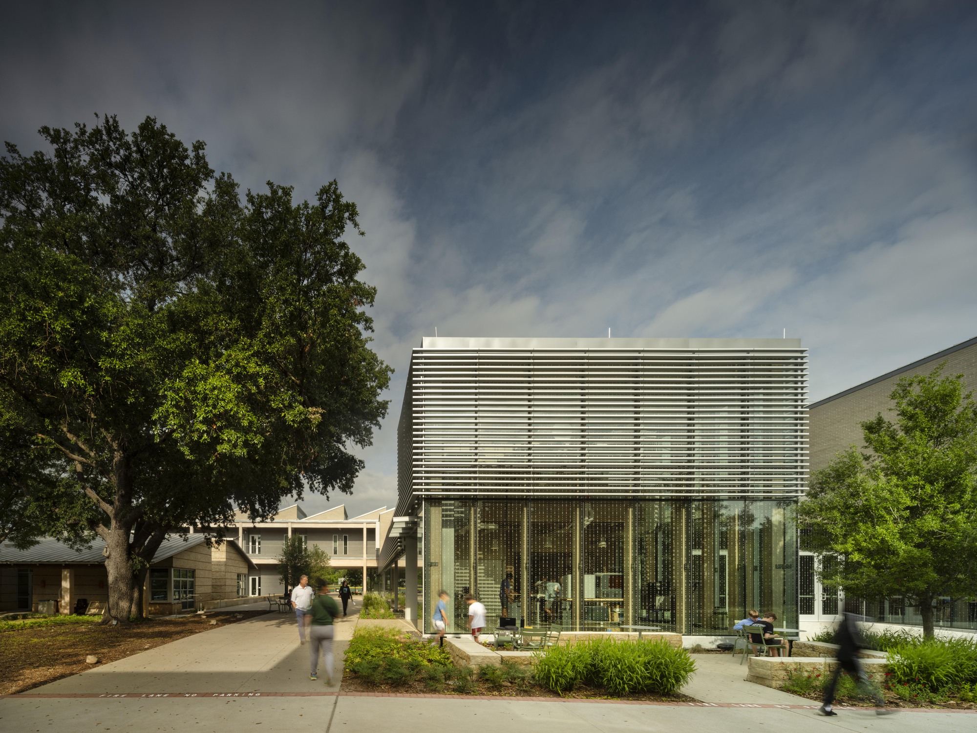 Glass box pavilion with horizontal sunshade louvers framed by a large tree and pedestrian pathway