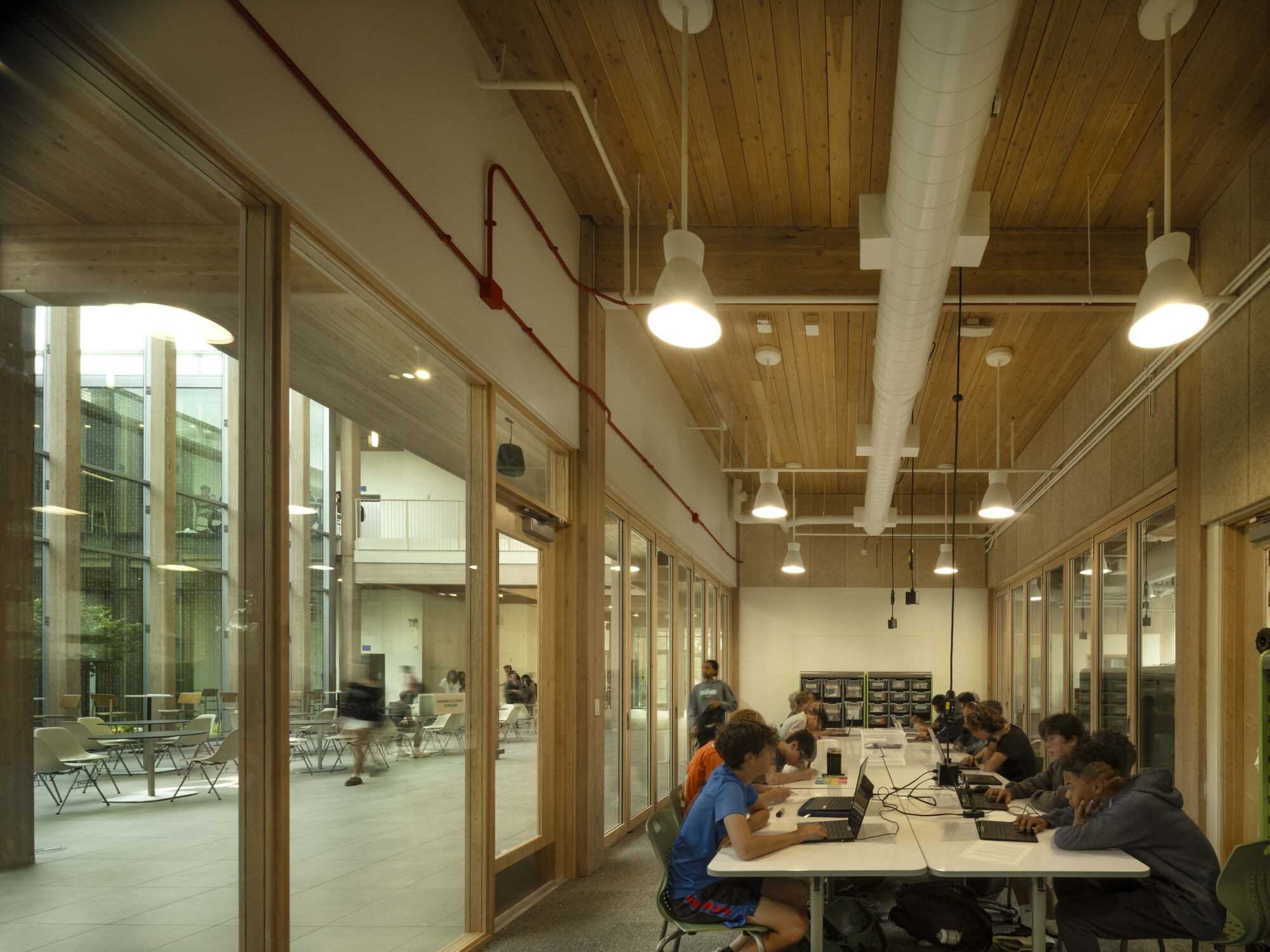 Study room with exposed timber ceiling, pendant lights and red conduit running along glass wall
