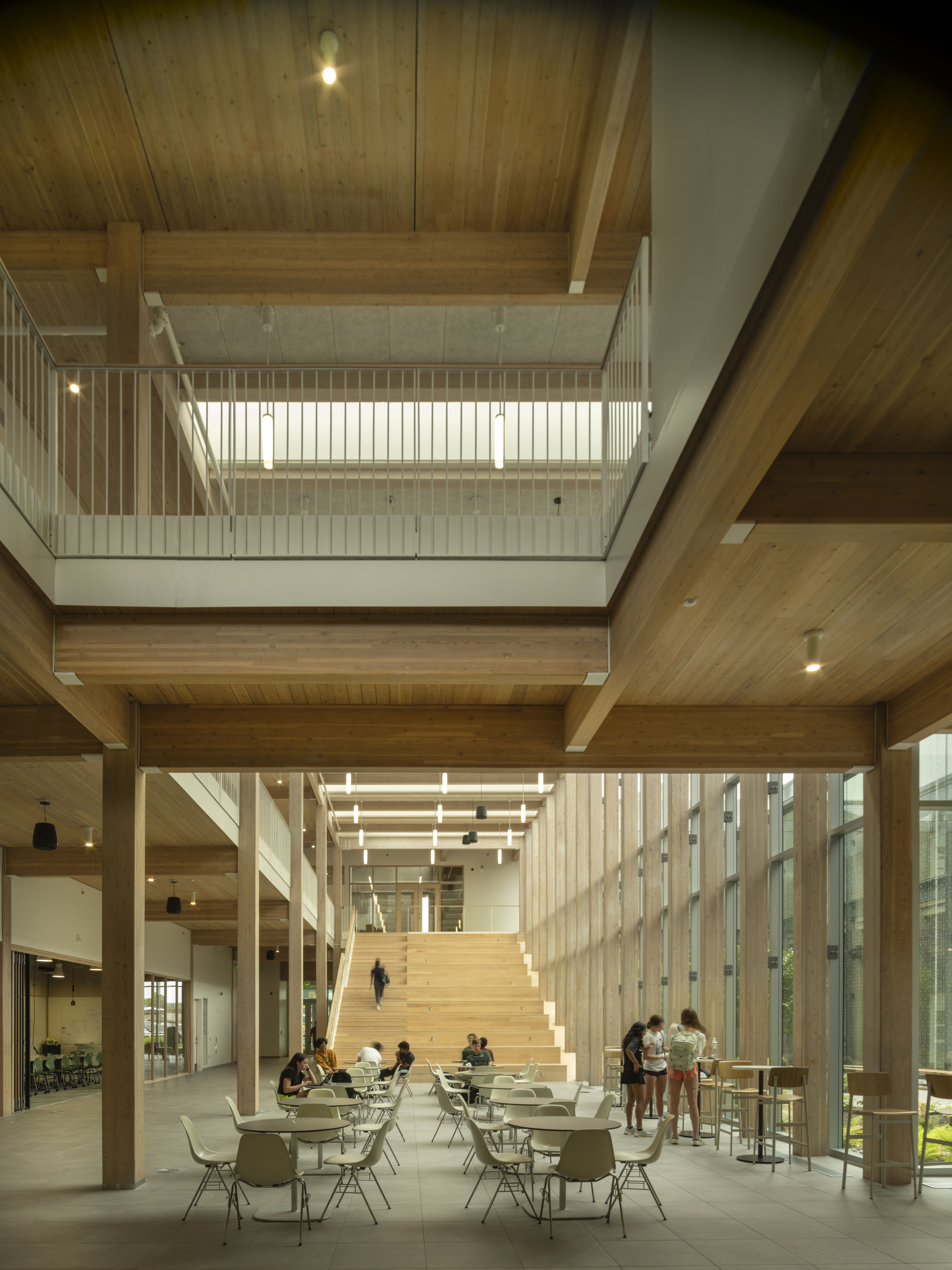 Triple-height atrium with timber beams, clerestory windows and wide staircase with people seated at tables below