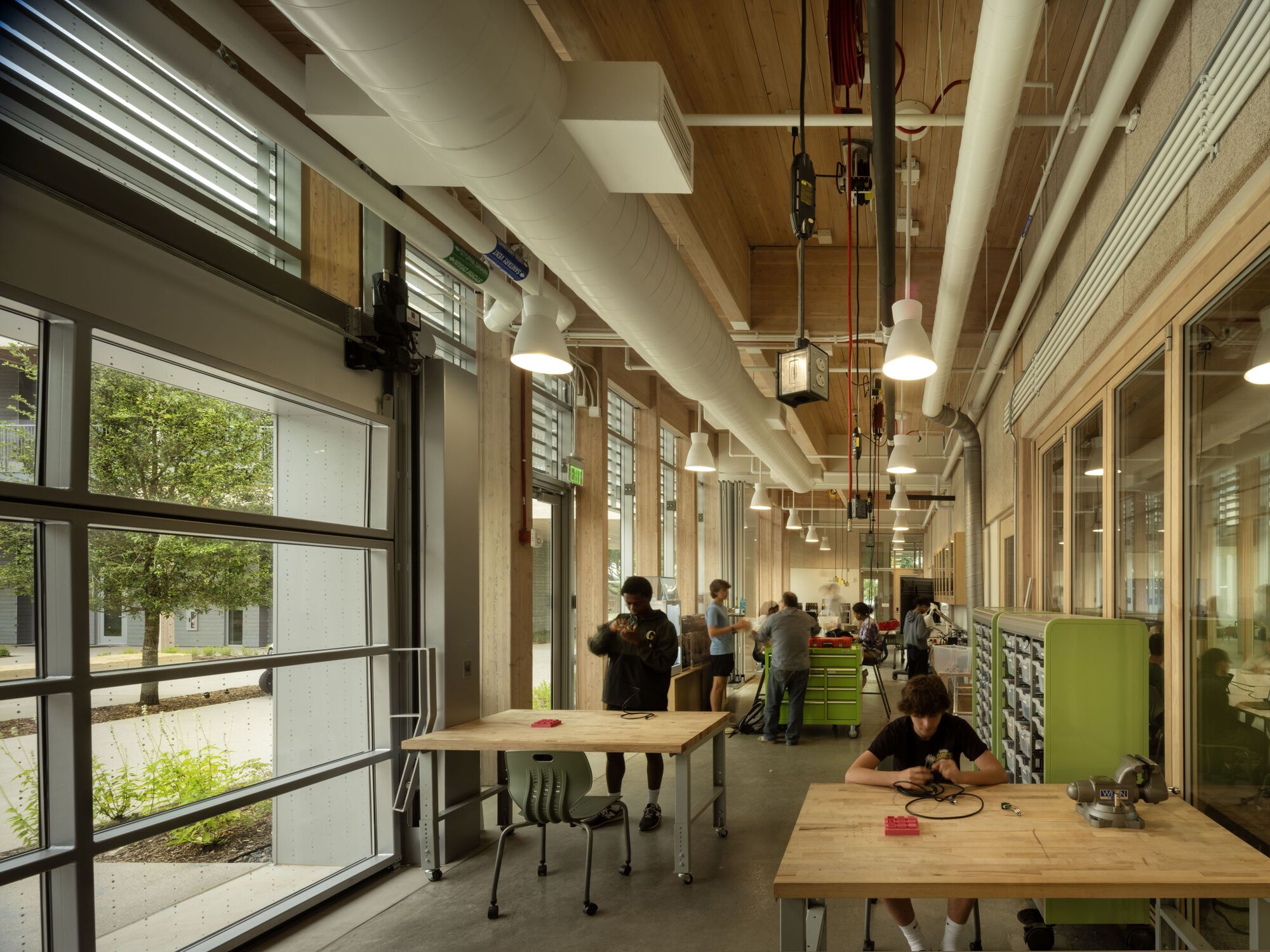 Workshop corridor with exposed timber ceiling, pendant lights, and workbenches along glazed walls with courtyard views