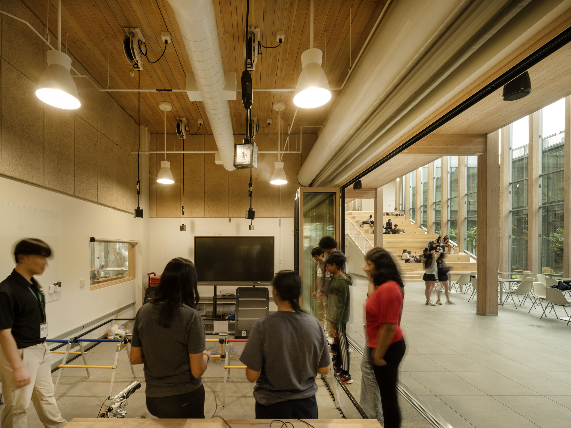 Glass-walled classroom space with timber ceiling and people gathered near a presentation screen