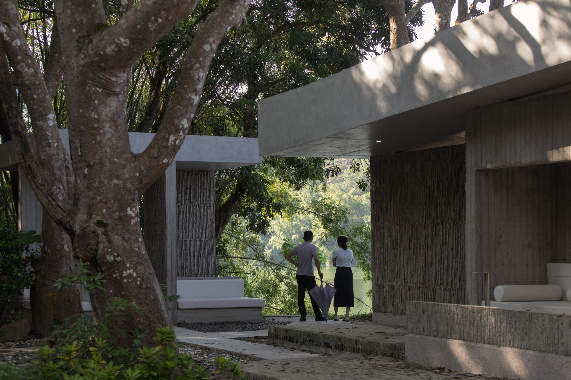 Two figures standing on a covered terrace framed by concrete beams and mature tree canopy