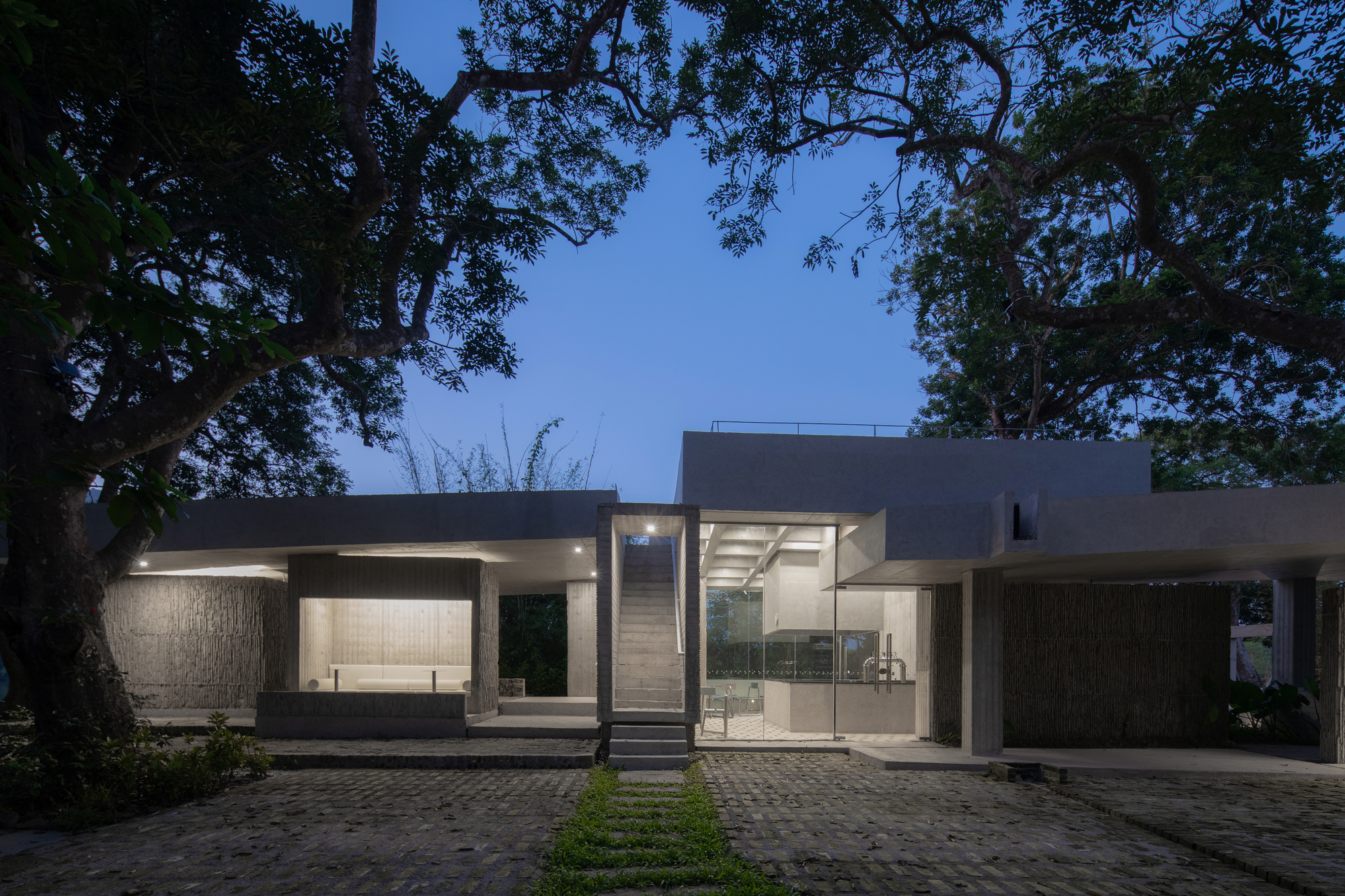Front facade at dusk with illuminated interiors beneath overhanging trees and cobblestone paving