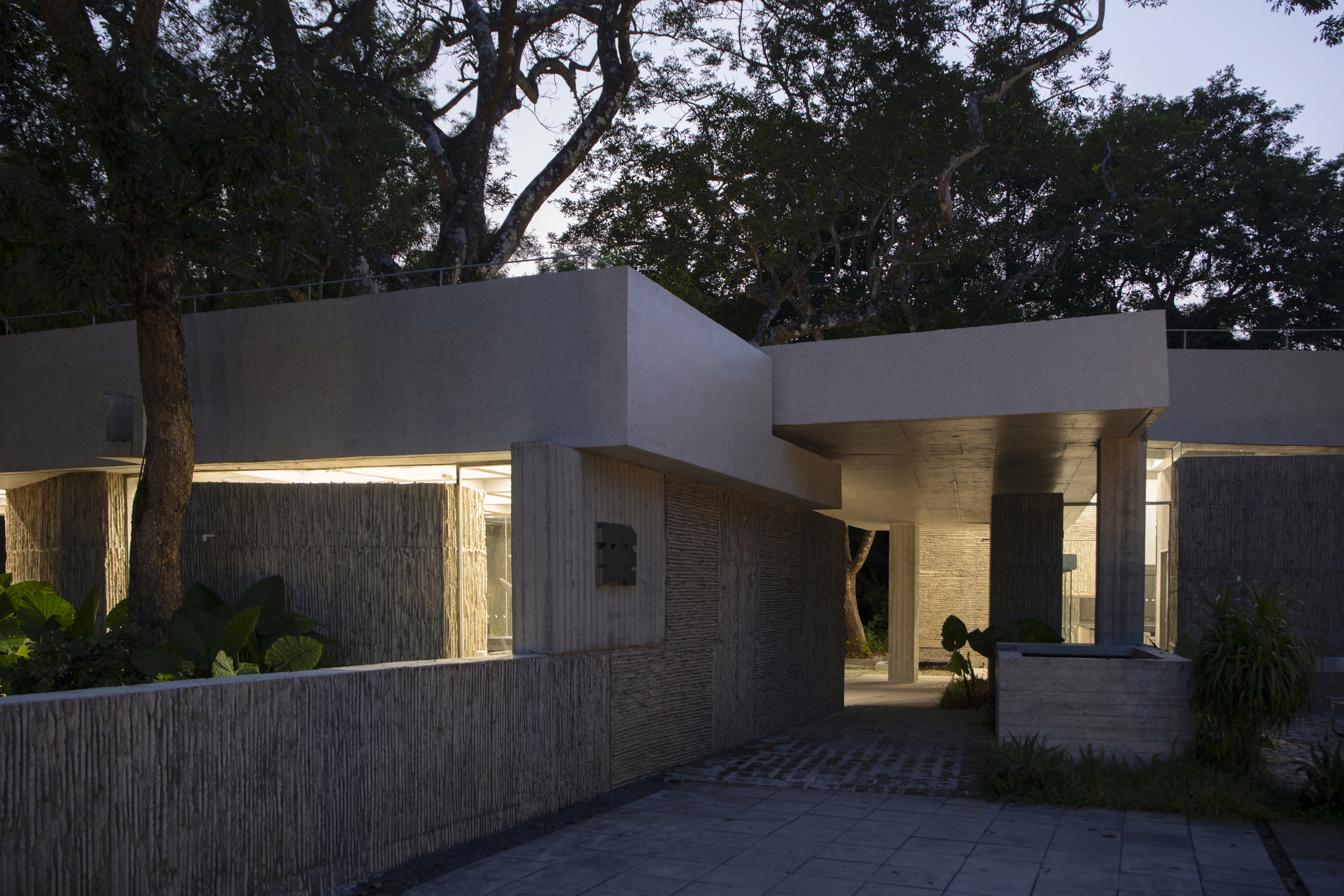 Entry courtyard with illuminated overhangs and vertical timber walls at dusk among tropical vegetation
