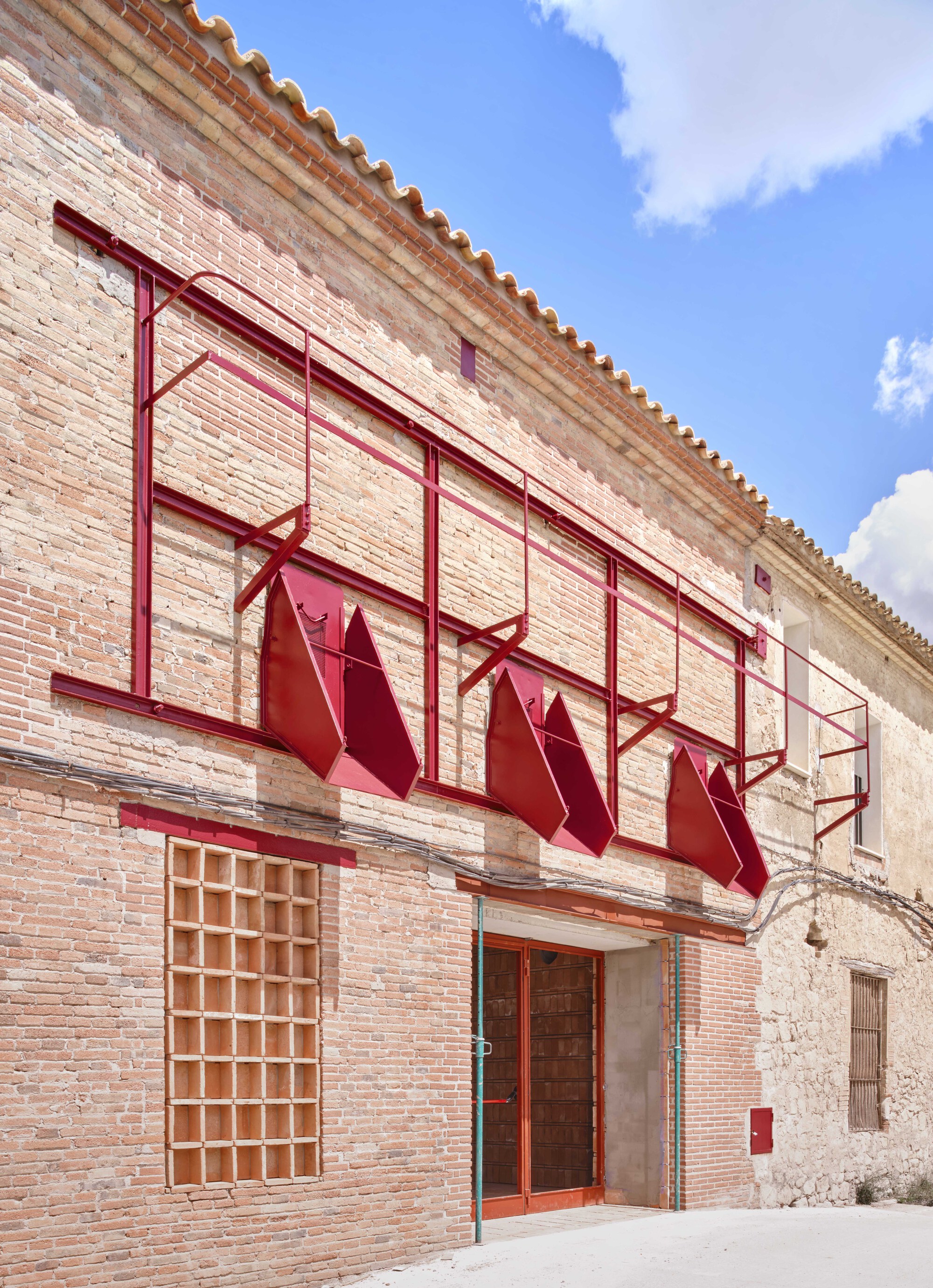 Brick facade with red metal awning frames and folded panels under a terracotta tile roof