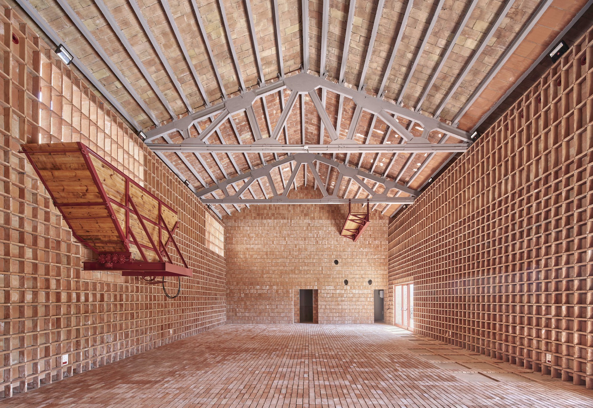 Interior hall with perforated terracotta brick walls, exposed timber trusses and a red steel balcony under diffused daylight
