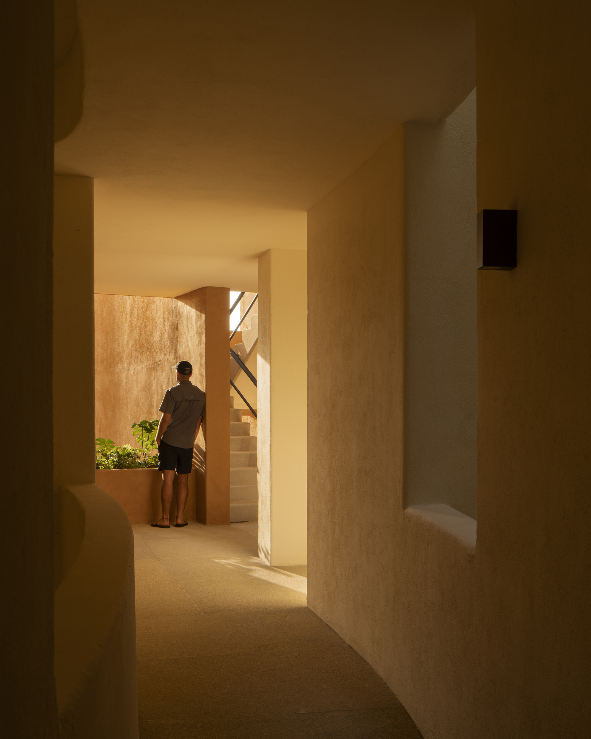 Interior passage with arched openings and warm light revealing a figure standing near a planted courtyard