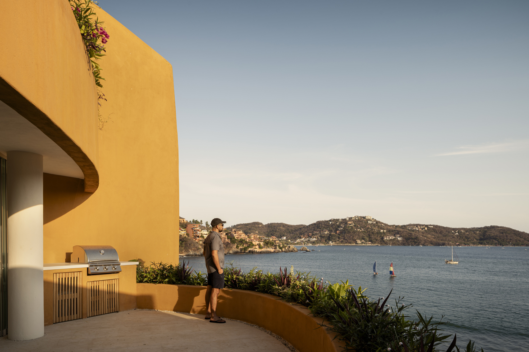 Rooftop terrace with outdoor kitchen overlooking a bay dotted with sailboats and hillside homes at dusk