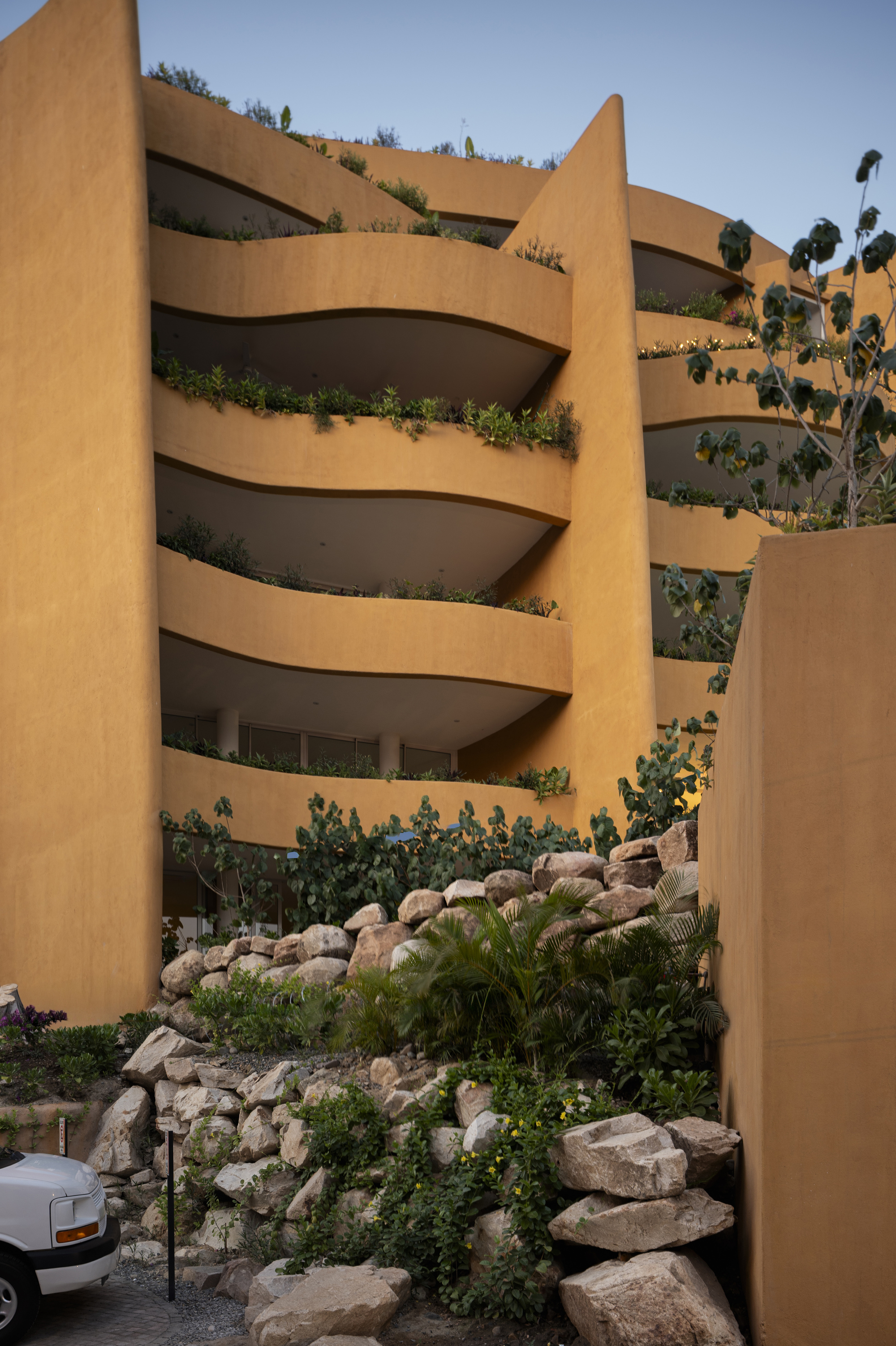 Planted balconies cascade down the ochre facade framed by a boulder retaining wall