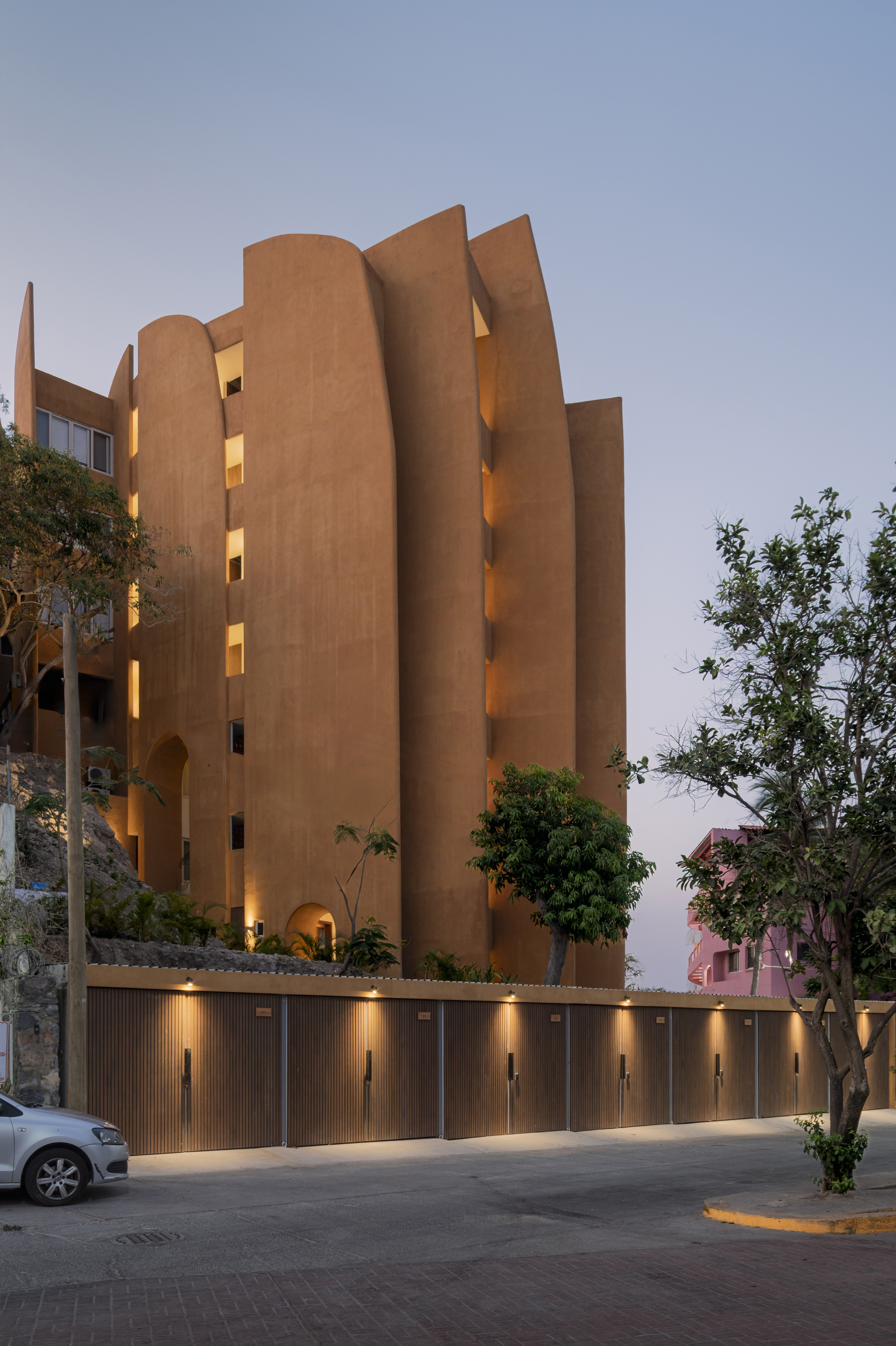 Curved ochre facade with illuminated balconies and a ribbed ground-level wall at twilight