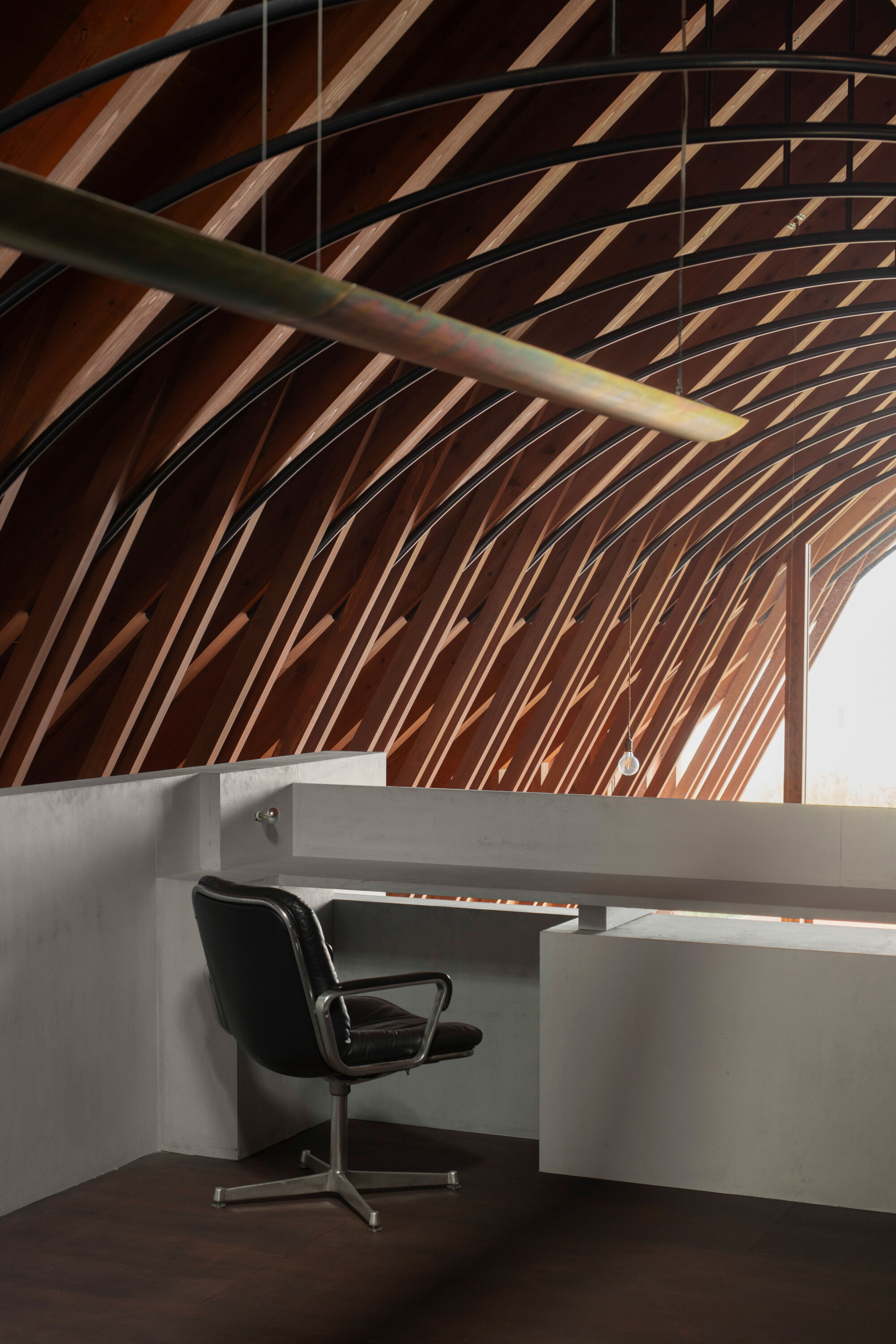 Built-in concrete desk with black office chair beneath curving timber ribs and green steel beam