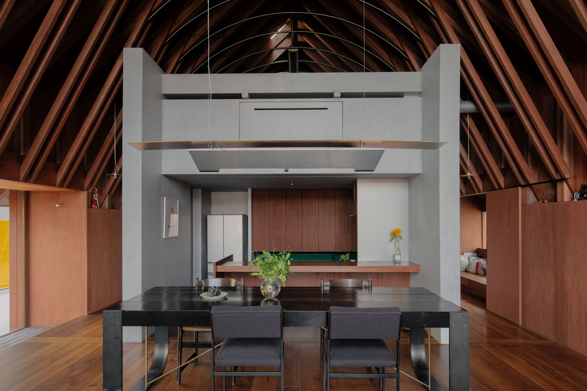 Interior view showing a freestanding grey volume beneath exposed timber roof rafters with dining table in foreground