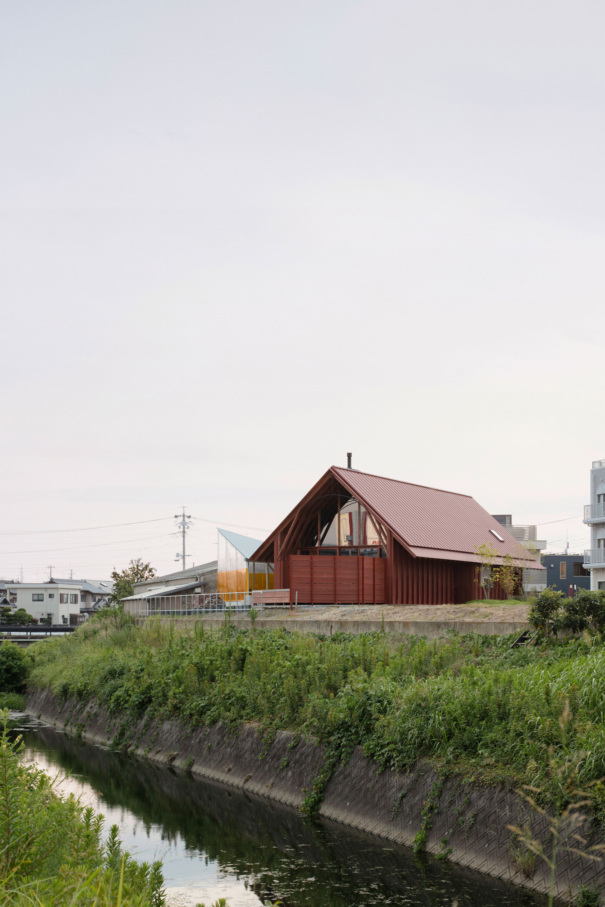 Gabled structure with pink metal roof and vertical timber cladding beside a canal under an overcast sky