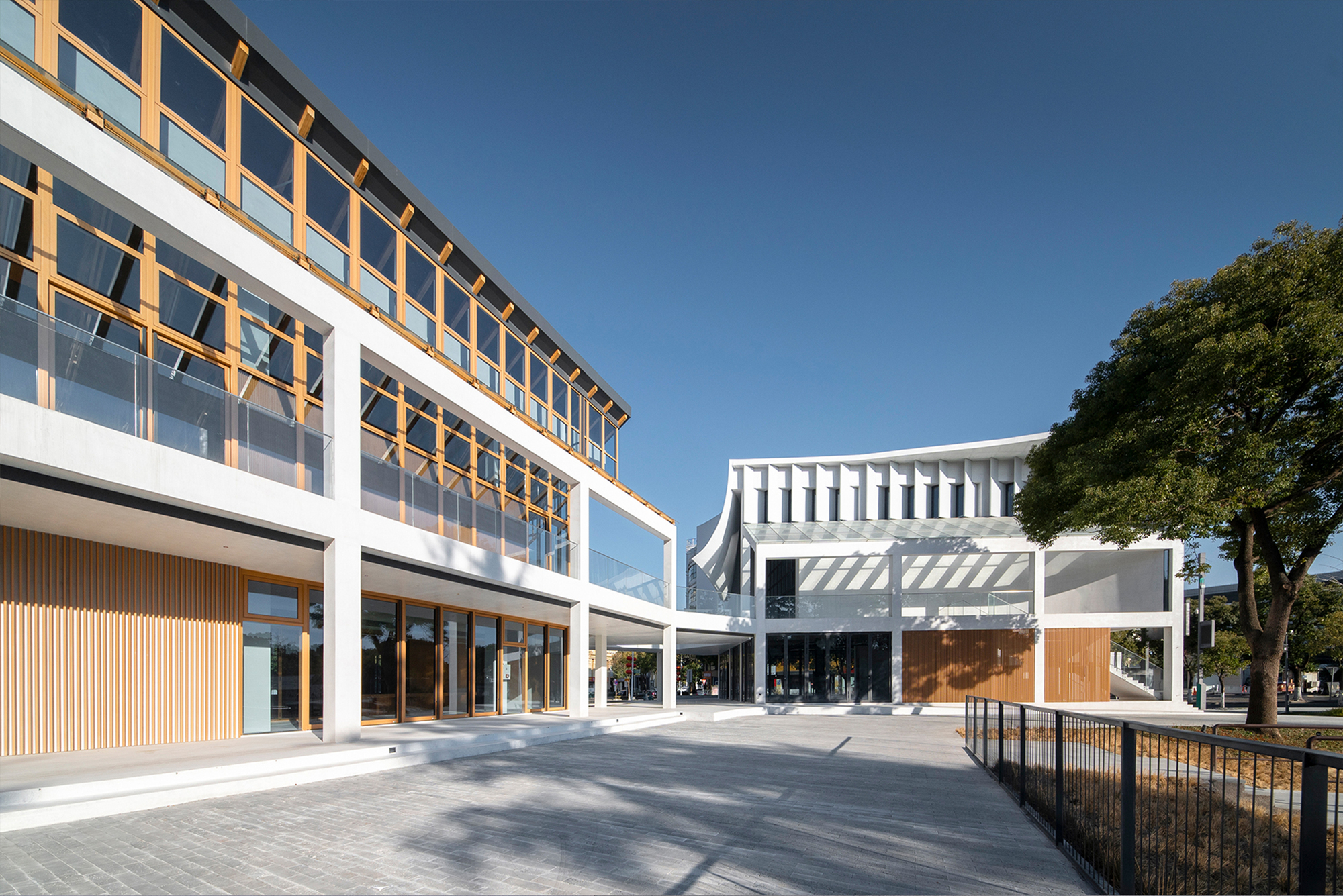 Courtyard between glazed timber frame building and white ribbed volume under clear sky