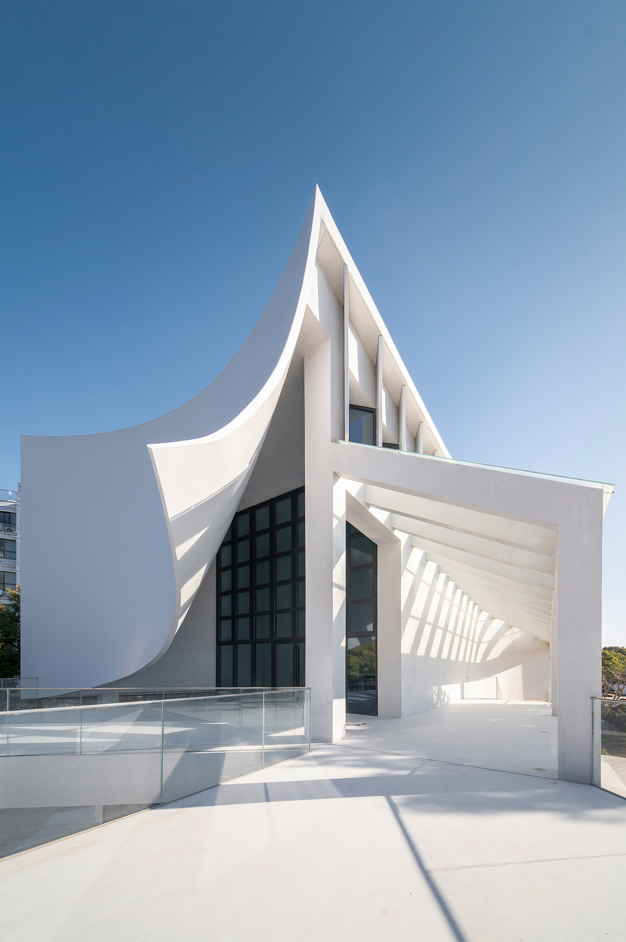White sculptural entrance with pointed canopy and folded planes framing a glazed opening under clear sky