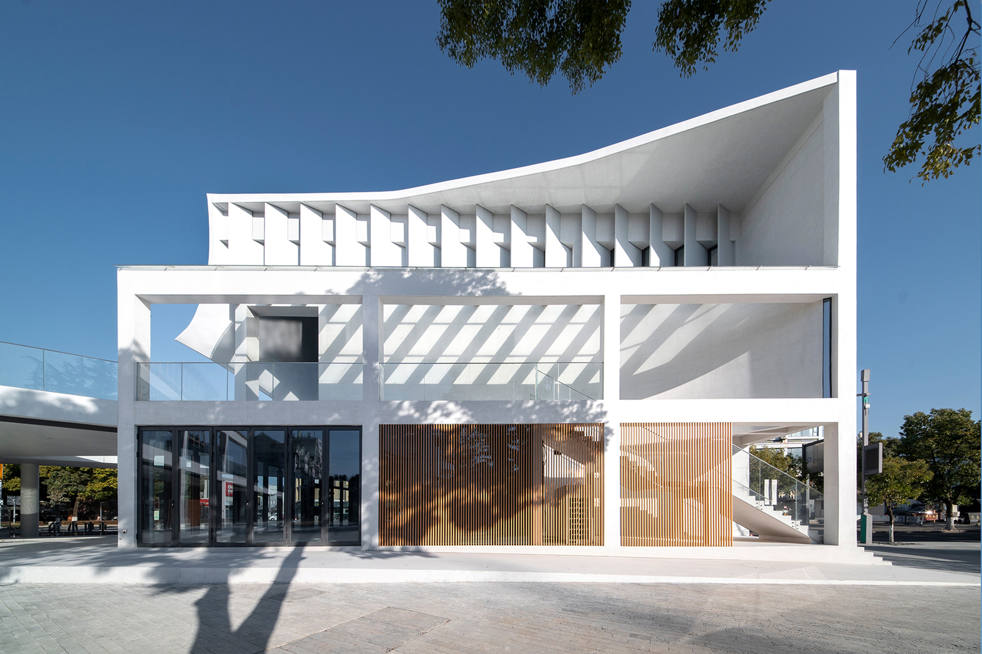 Street elevation showing layered white volumes with recessed timber screens and deep shadow patterns in afternoon light