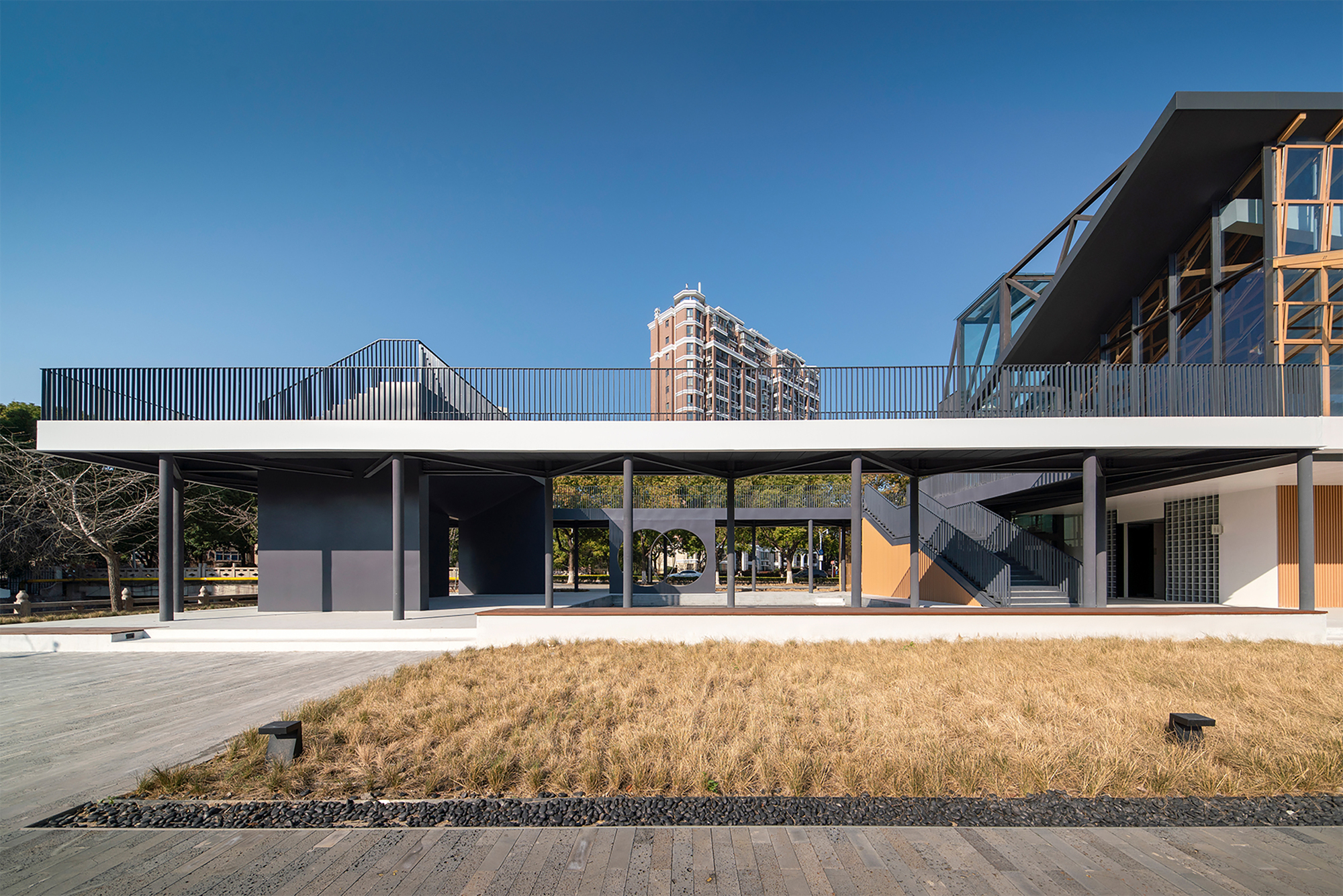 Elevated walkway with vertical screening connecting two volumes above a ground-level plaza in dry grass