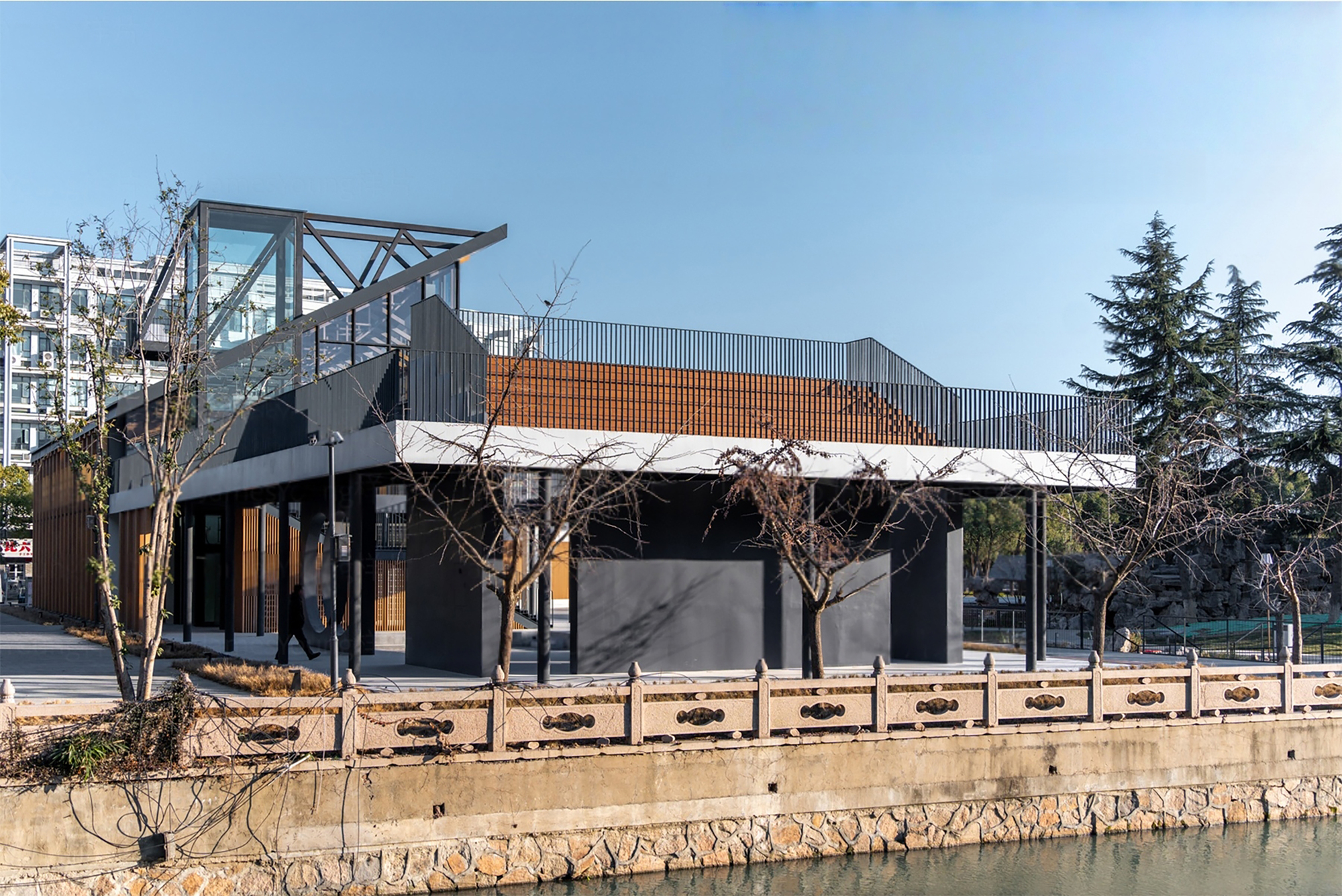 Waterside view of the corrugated metal roof and exposed steel truss above dark facades and bare winter trees