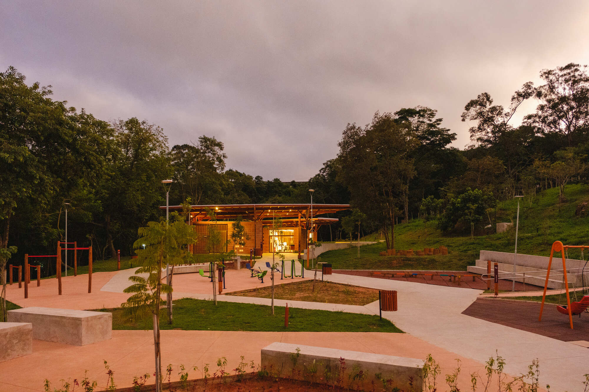 Community building with timber roof overlooking a plaza with young plantings and playground equipment at twilight
