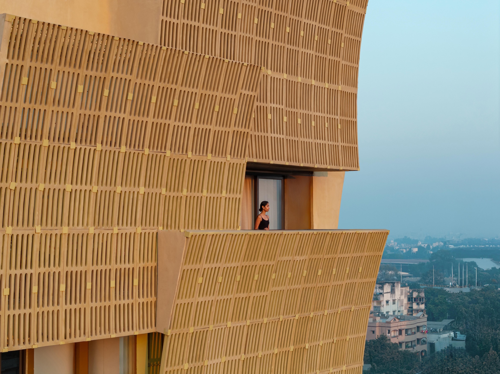 Person standing on a cantilevered balcony framed by ribbed brick screens overlooking the distant cityscape at dusk