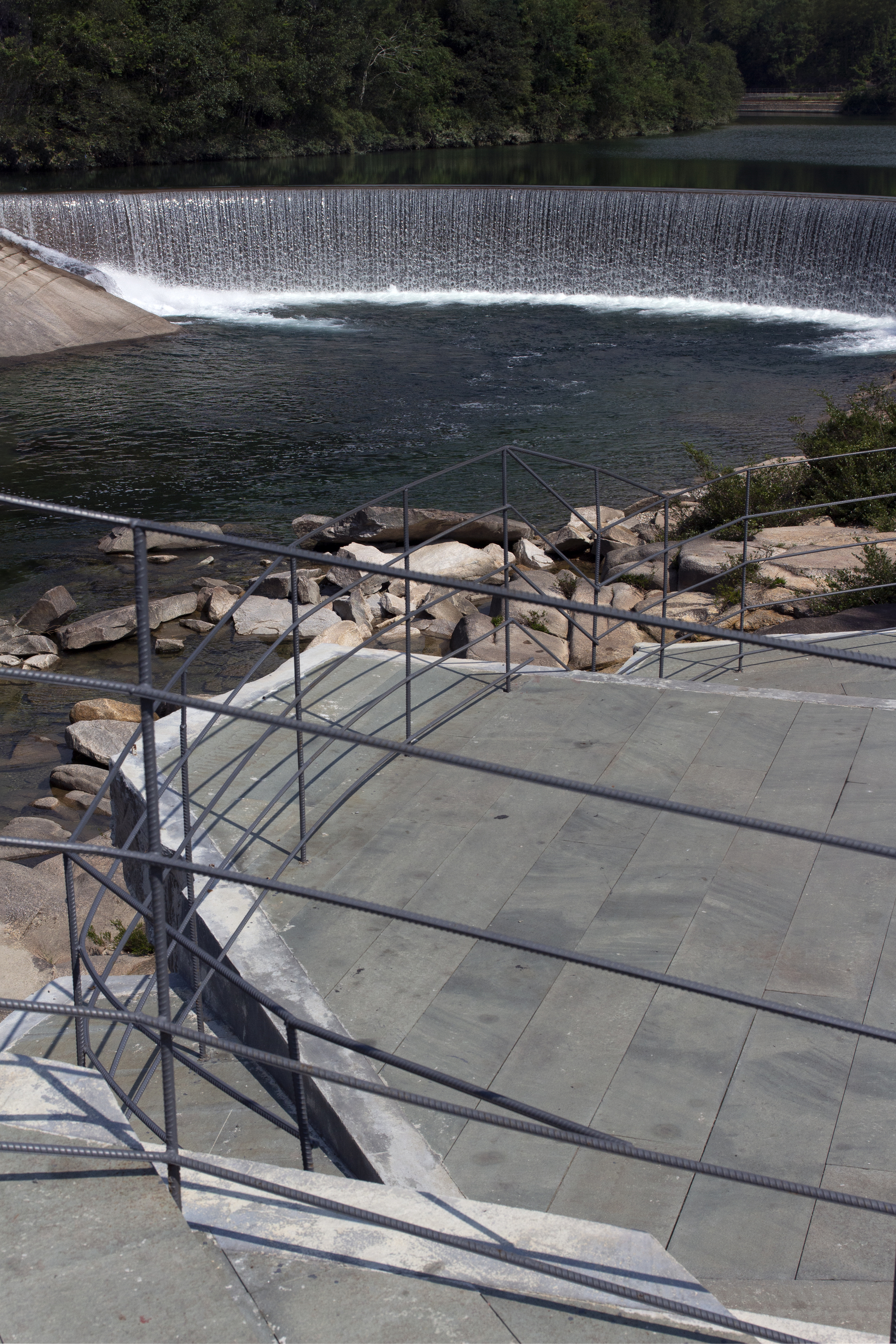 Stone terrace with geometric metal railing overlooking the dam weir and forested hillside