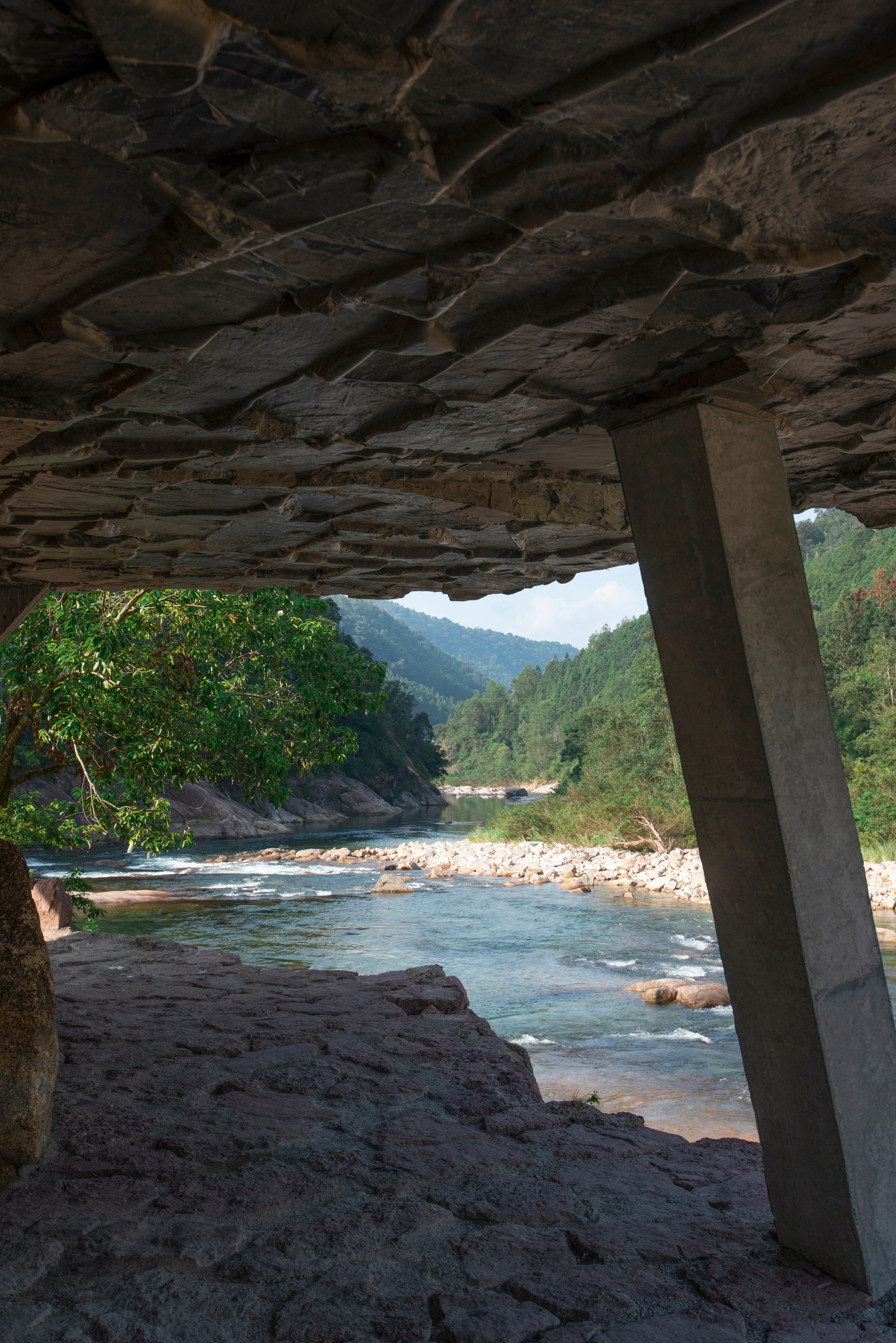 View from beneath a layered stone overhang toward a river winding through forested mountains