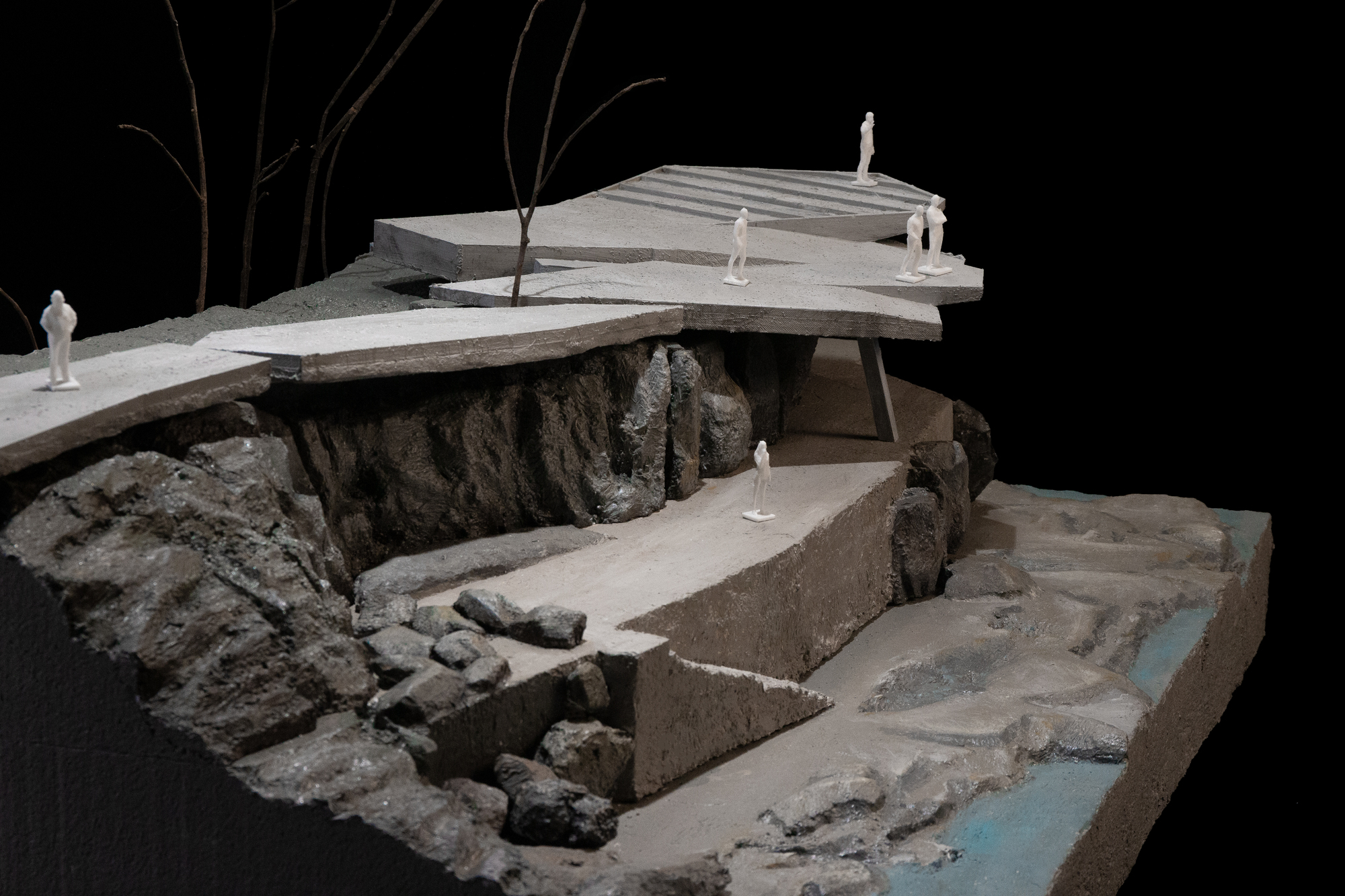 Physical model photographed against black backdrop showing cantilevered terraces over sculpted rocky riverbed with miniature figures