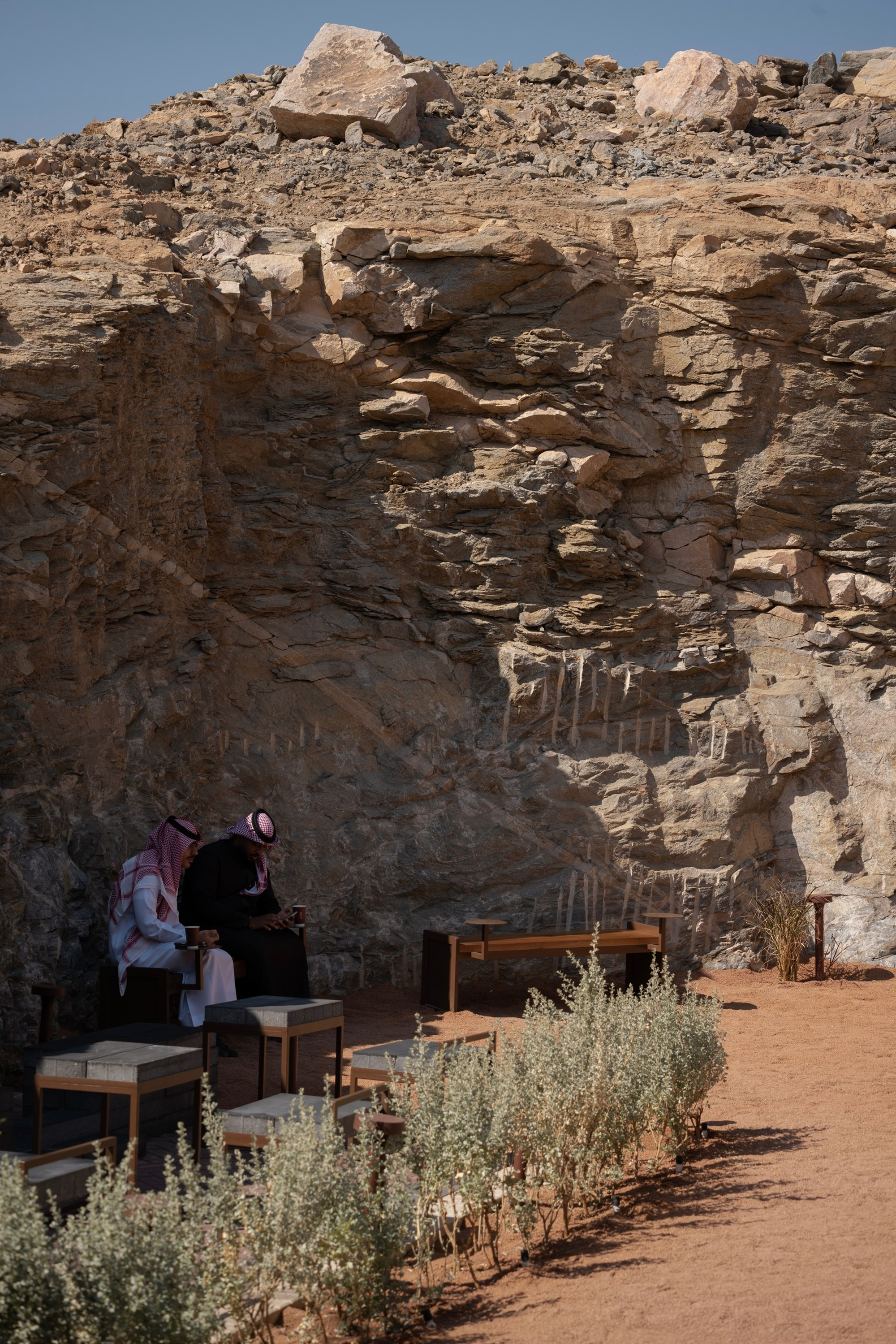 Wooden seating and planted beds at the base of a layered sandstone cliff under blue sky