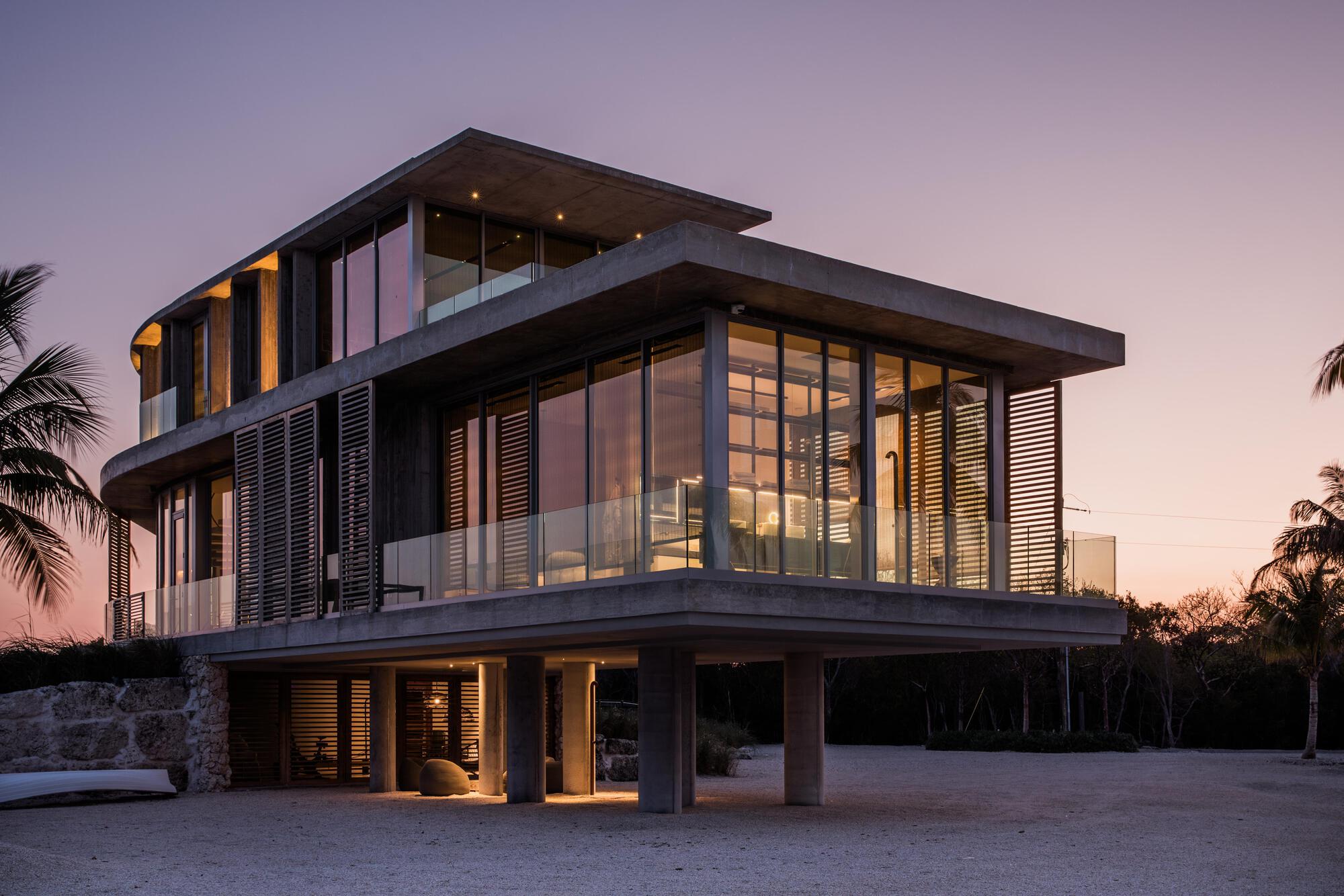 Corner view of the three-level residence with illuminated interiors at dusk among palm trees