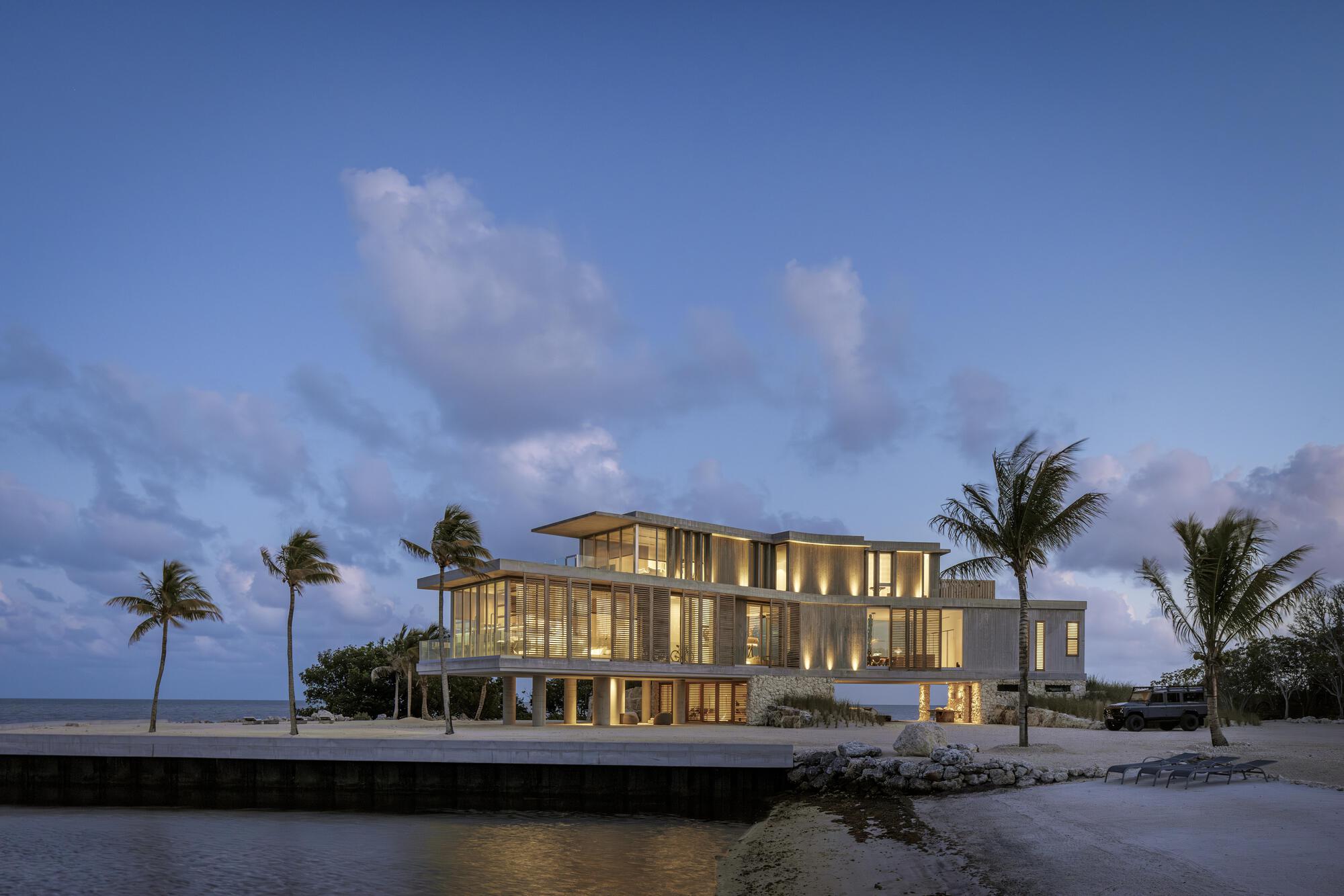 Waterside elevation showing the illuminated glass facades and concrete structure at twilight with swaying palms